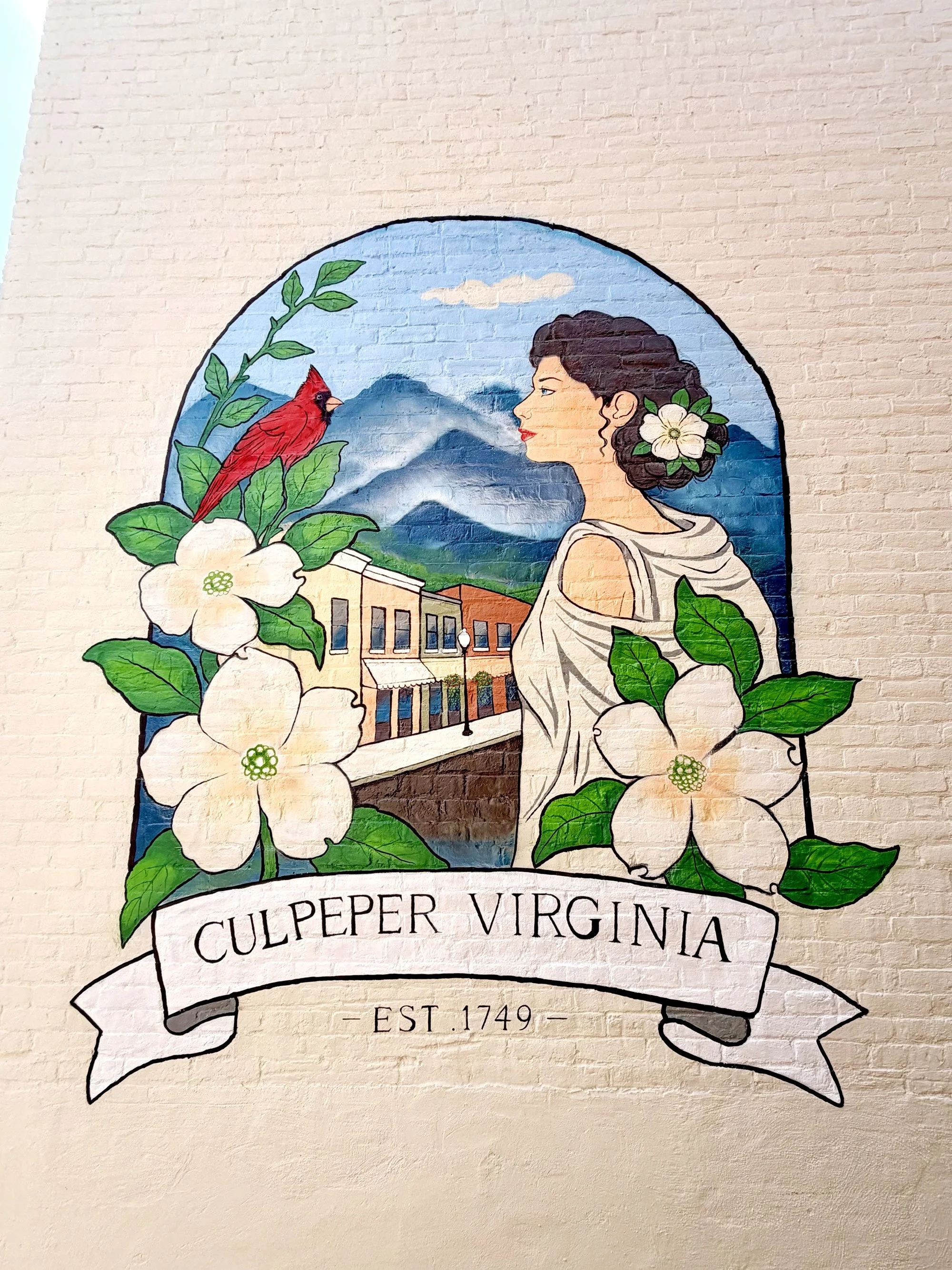 A mural with a drawing of a woman's profile with curly dark hair adorned with white flowers, a mountain landscape in the background, and a red bird perched on a branch with green leaves. Below is a banner with the text "Culpeper Virginia" and "Est. 1749".
