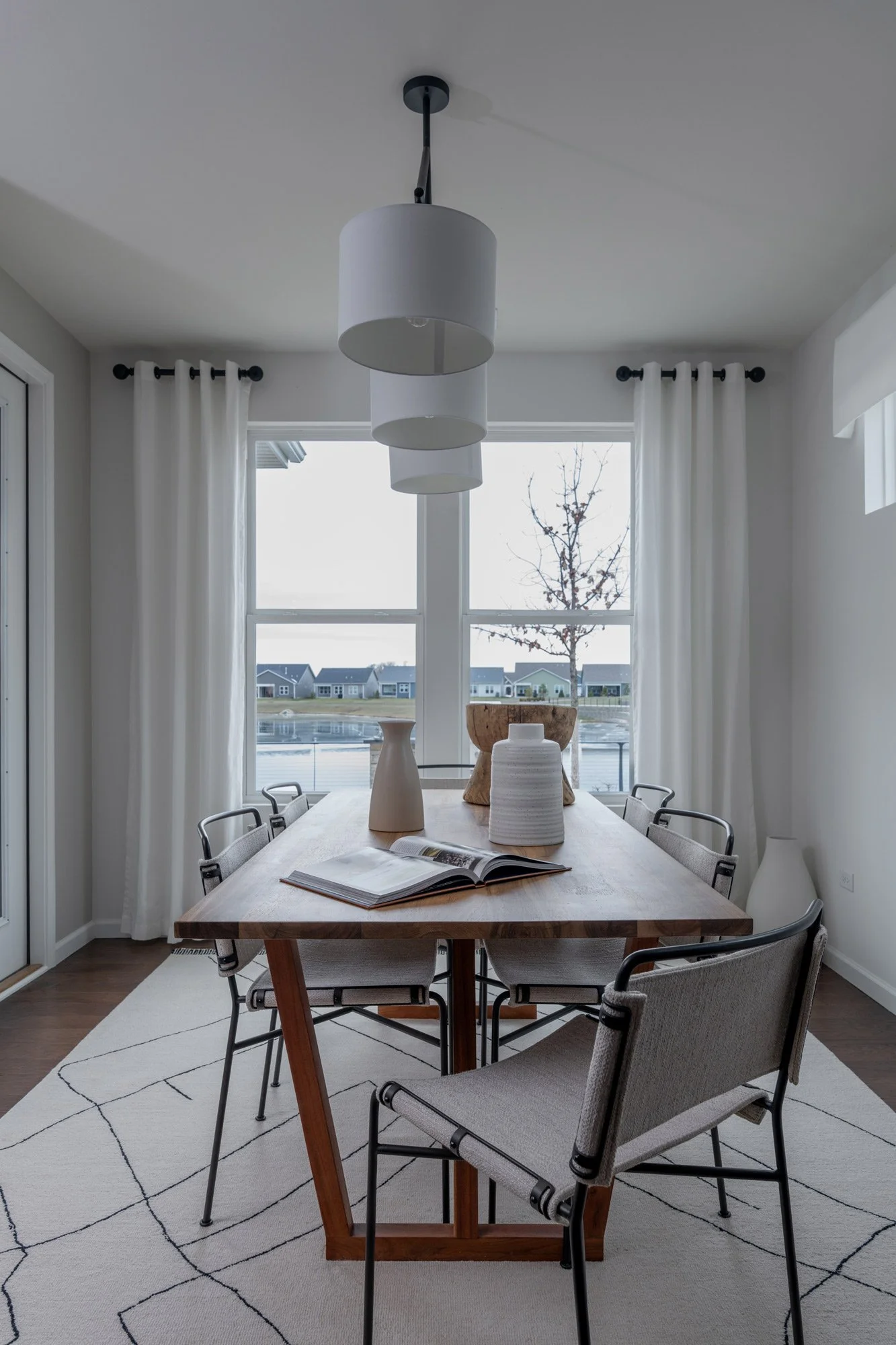 Modern dining room with a wooden table, six chairs, white curtains, and decorative vases on the table. Large window shows house and trees outside. Overhead light fixture with three white shades.