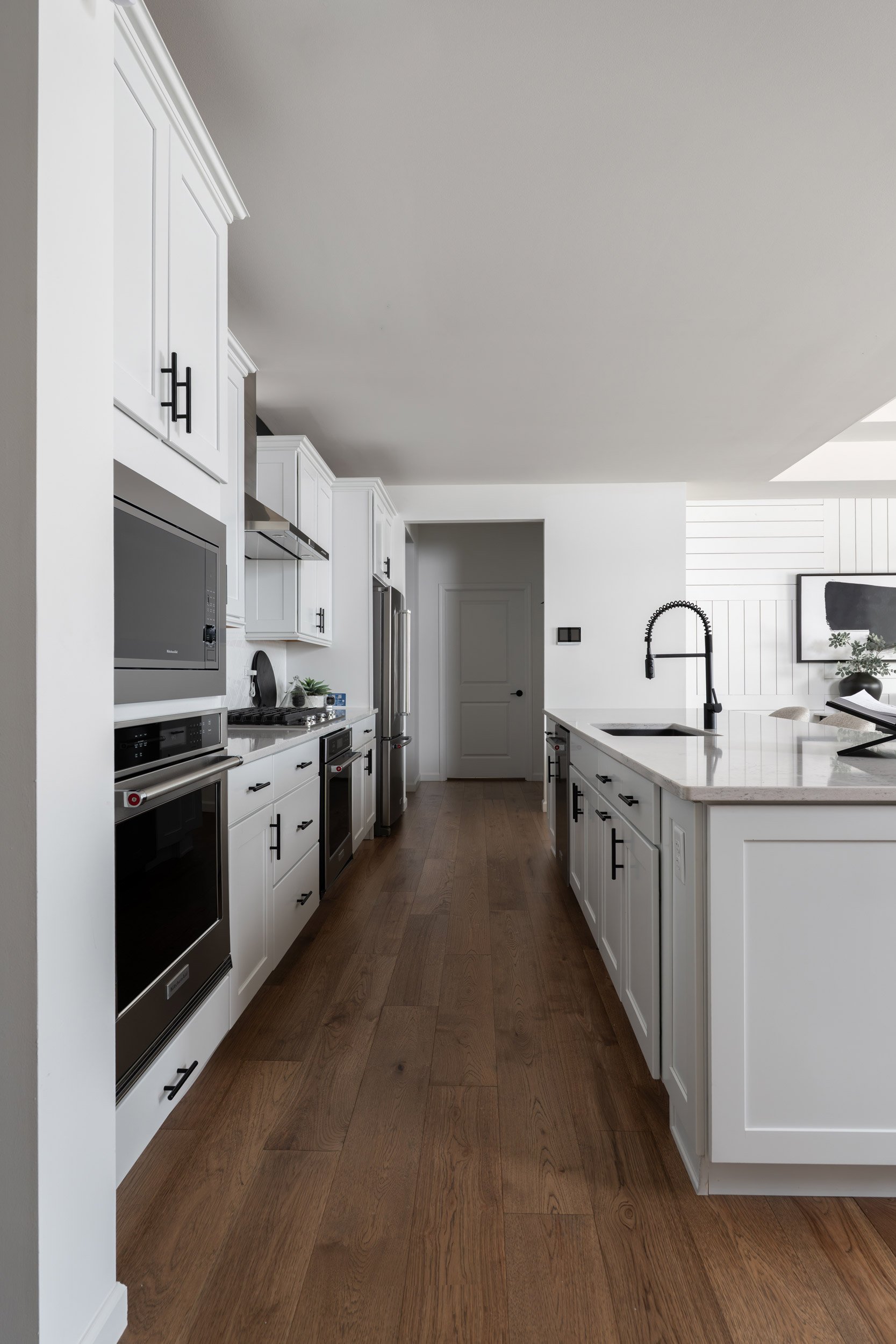A modern kitchen with white cabinetry, stainless steel appliances, and a wooden floor. There is a black faucet and a large kitchen island with a granite countertop.