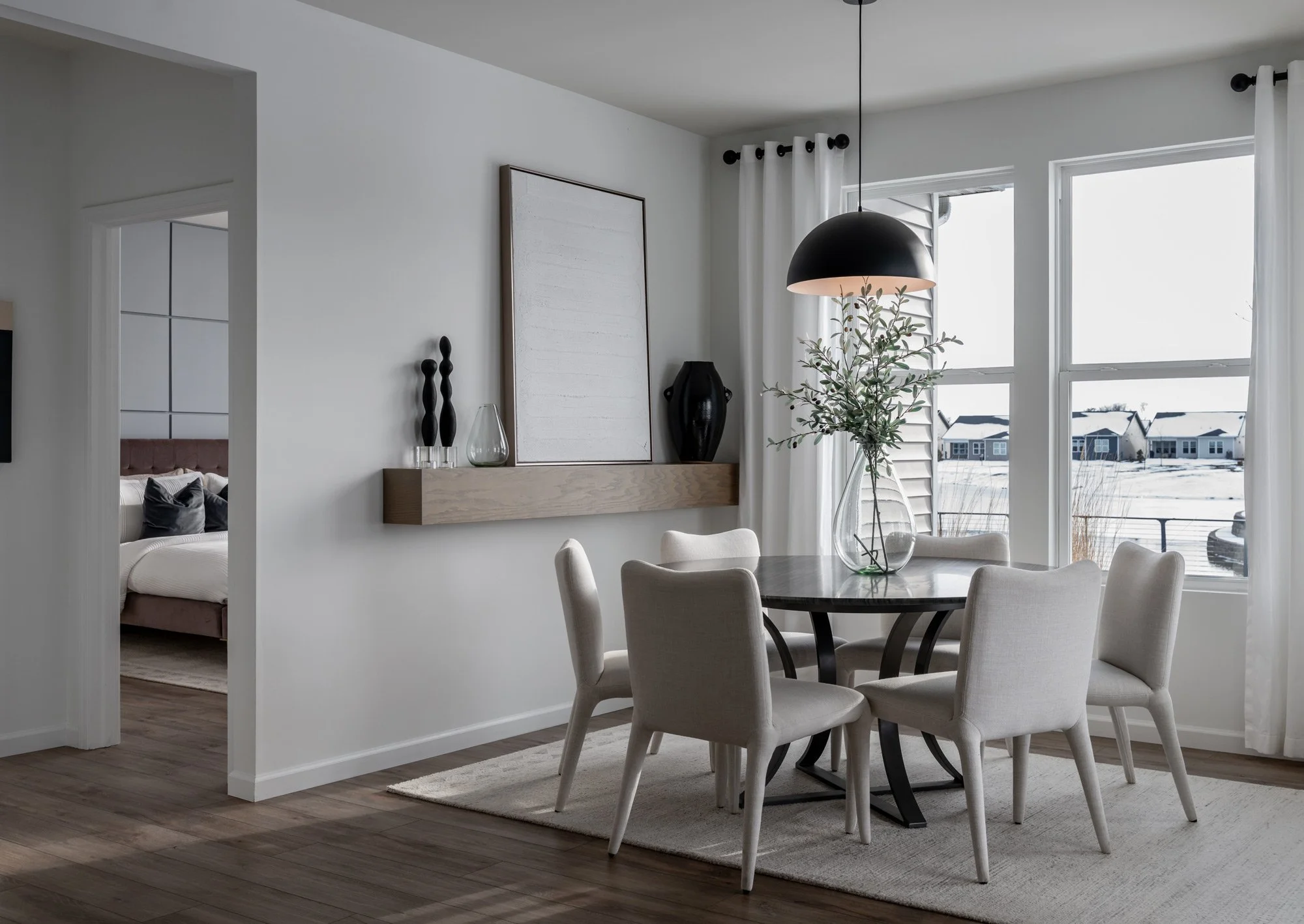 A dining room with a round dark table, six beige chairs, a large glass vase with green branches, large windows with white curtains, a modern pendant lamp, and a wooden shelf with decorative vases and artwork. A bedroom is visible through an open door