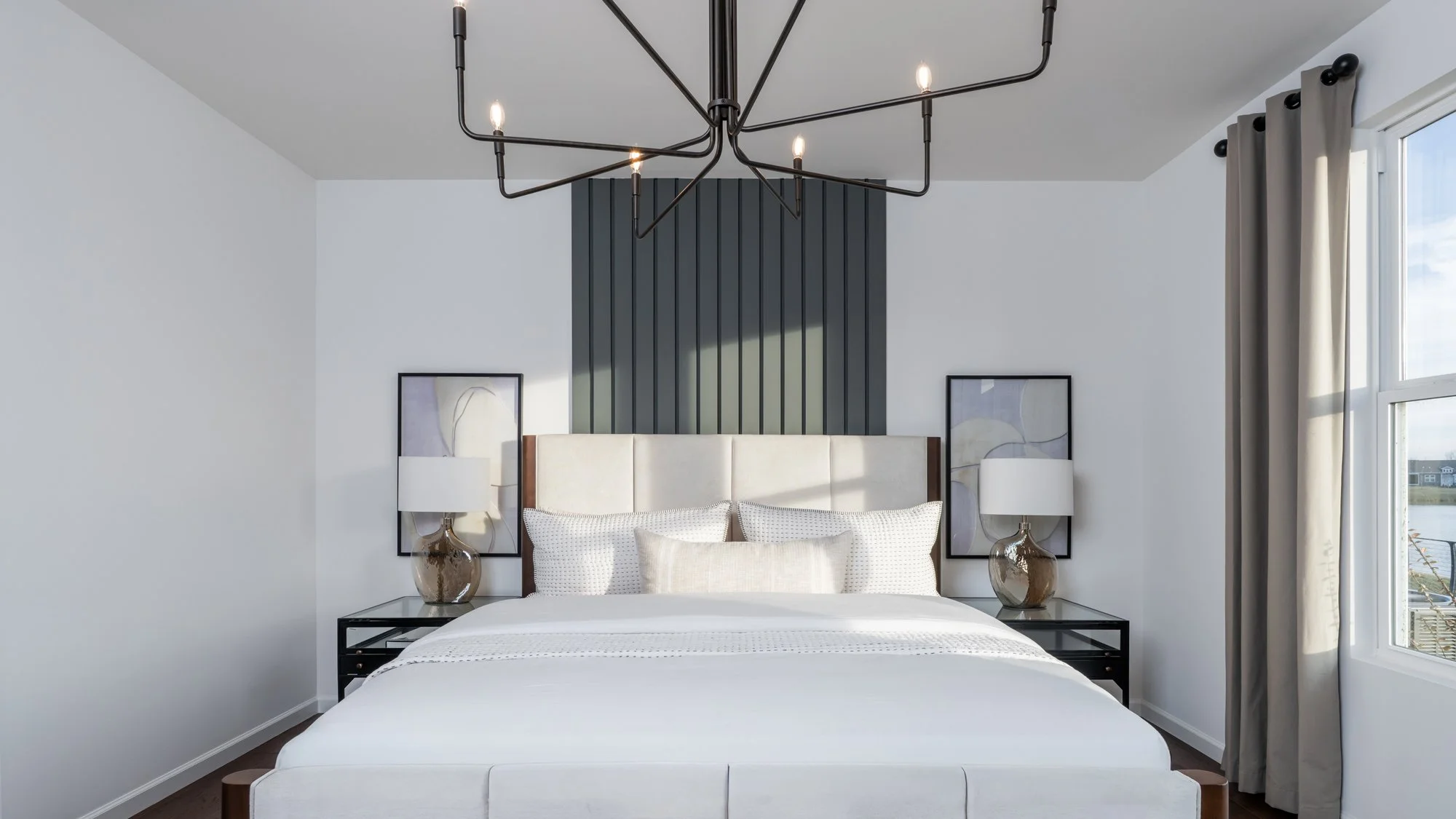 Modern bedroom with white walls, large window with gray curtains, black pendant light fixture, beige bed with white pillows, black bedside tables with glass tops, and artwork.