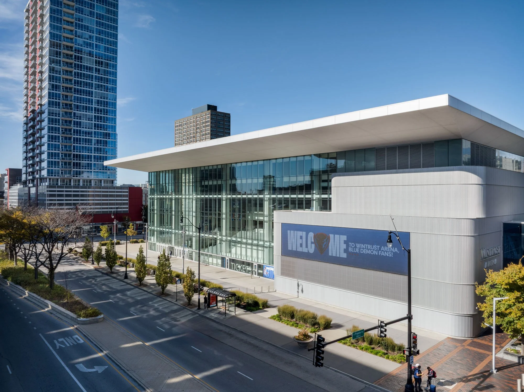 Modern arena building with glass facade and large electronic banner displaying welcome message to Wintrust Arena for Blue Demon fans. Street in foreground with trees, bus stop, and traffic lights, surrounded by tall buildings under a clear blue sky.