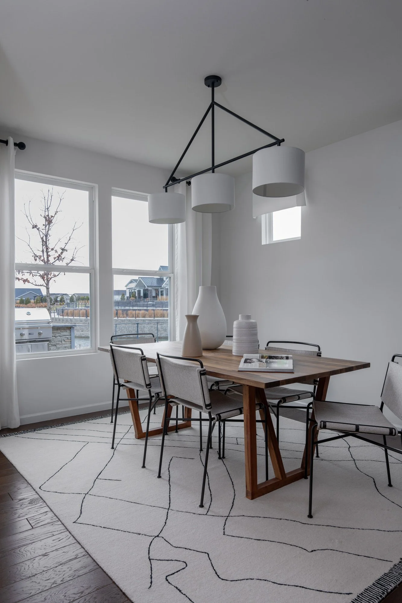 Dining room with a wooden table, six chairs, white vases, and a black and white abstract patterned rug, illuminated by a modern chandelier, with large windows showing a suburban neighborhood outside.