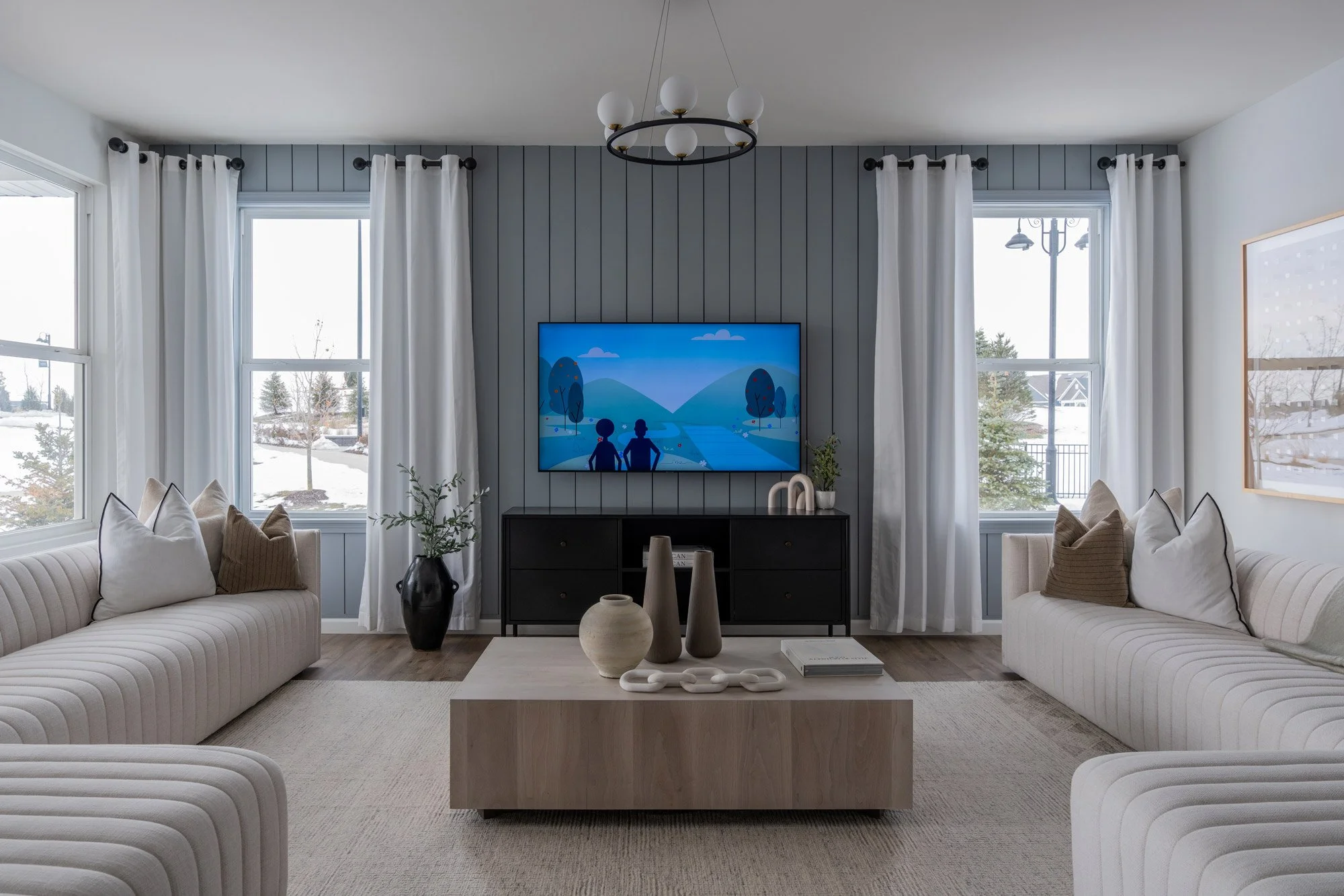 Living room with two beige striped sofas, a wooden coffee table with vases and books, a black TV stand with a mounted TV, large windows with white curtains, and a modern chandelier.