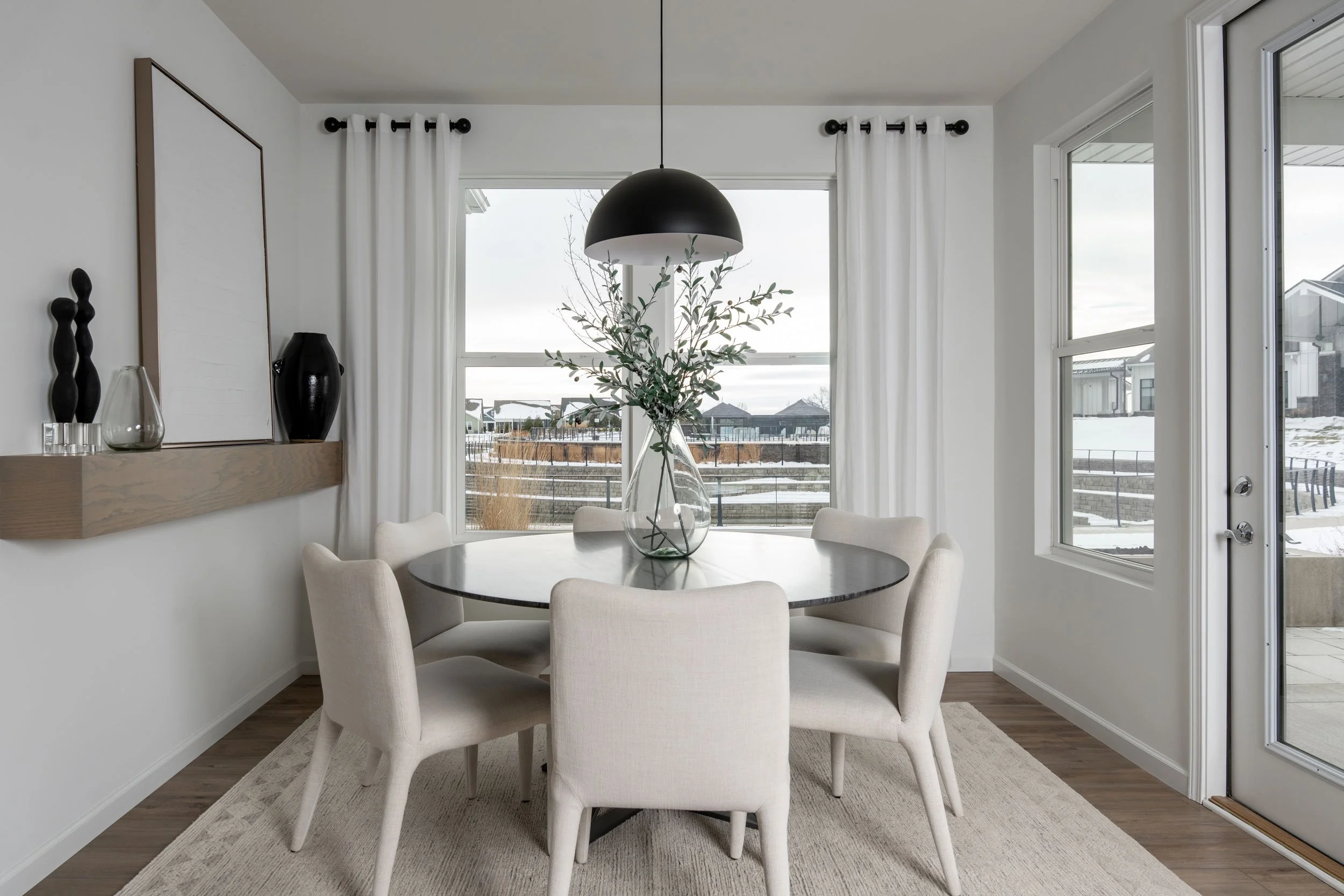 Modern dining room with a round table, six white chairs, a vase with green foliage as a centerpiece, large windows with white curtains, a wooden shelf with decor, and a door leading outside, overlooking a snowy landscape.