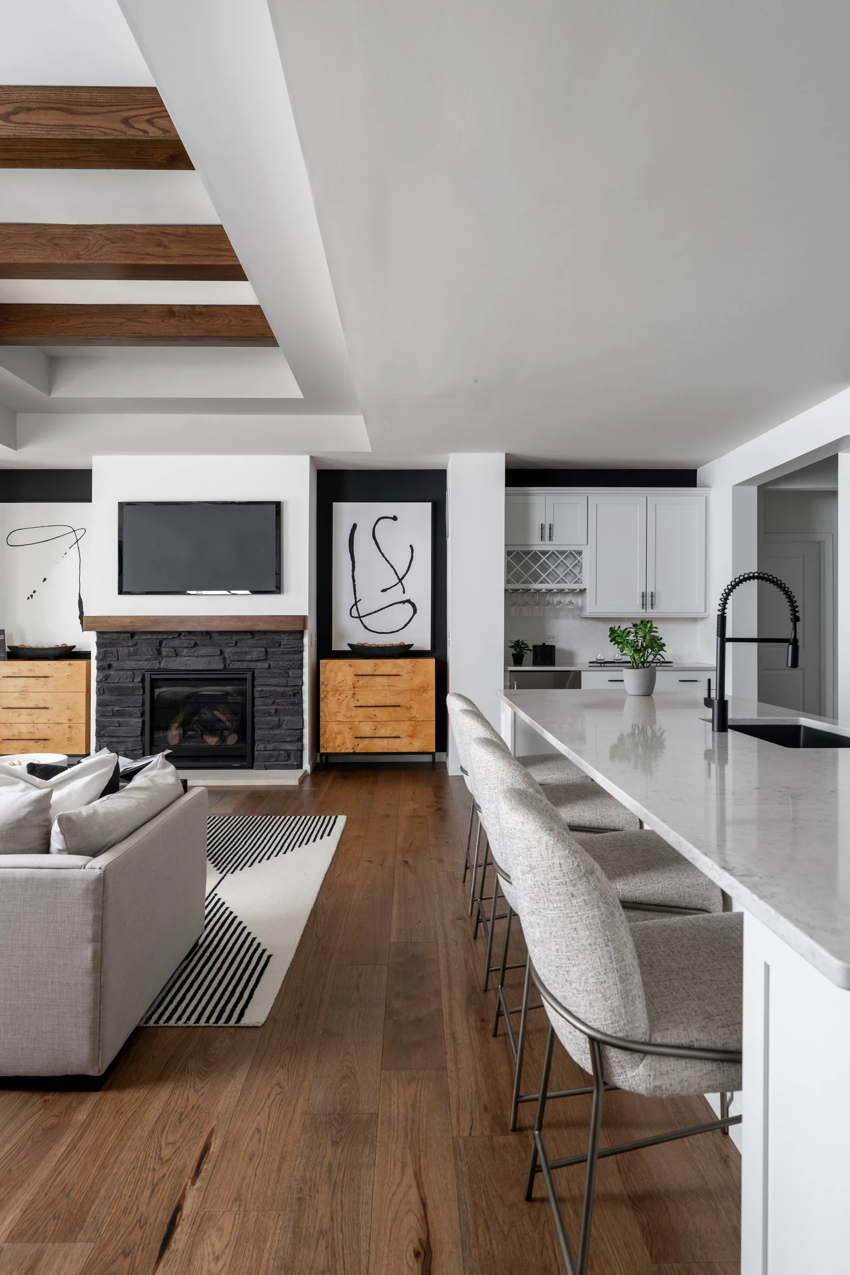 Modern living room and kitchen with white walls, wooden ceiling beams, a fireplace with a black stone surround, wall-mounted TV, white cabinetry, a marble kitchen island with barstools, and minimalistic decor.