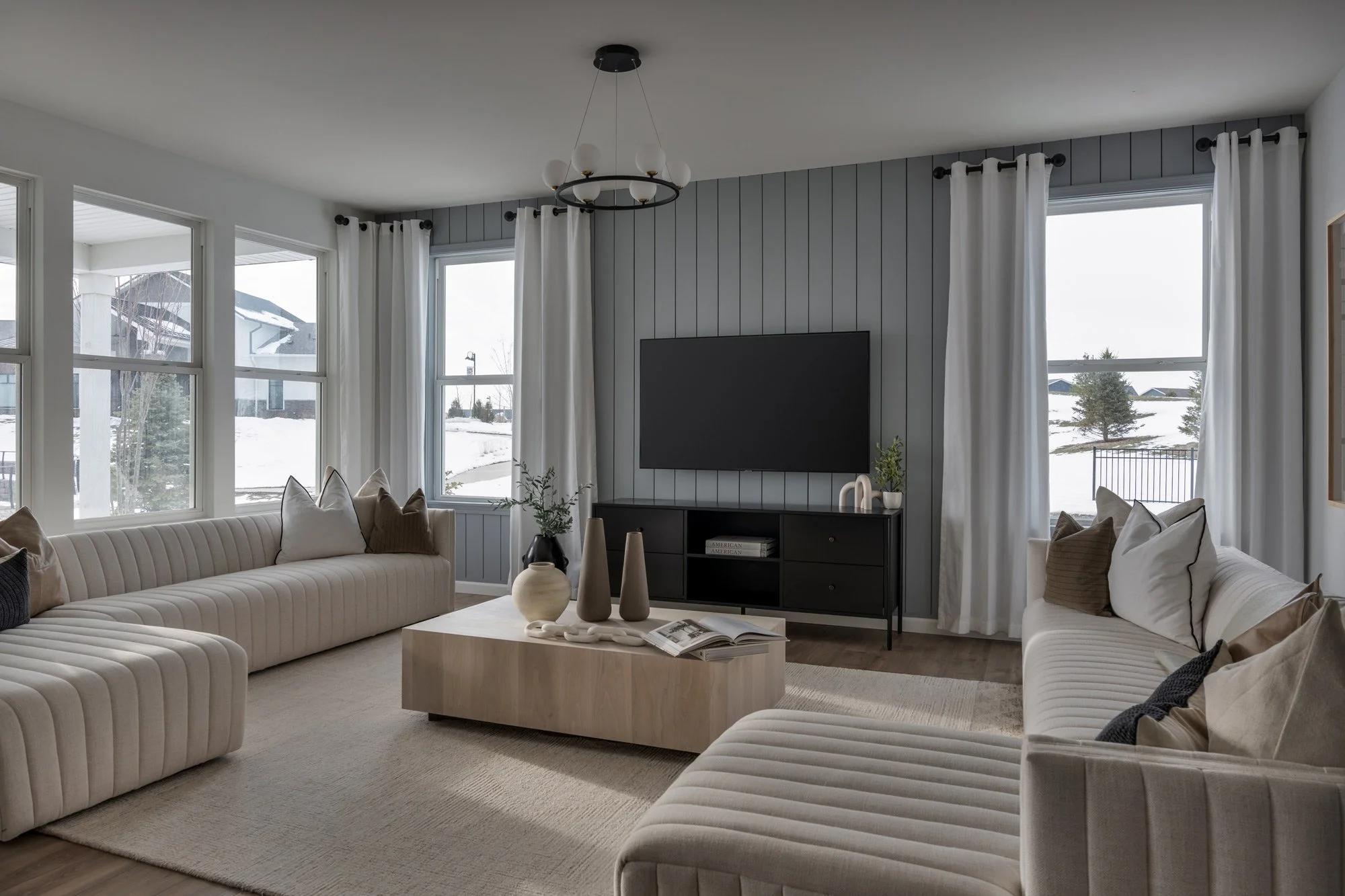 Living room with two white sofas, a light wood coffee table, decorative vases, and a TV on a gray accent wall, with large windows showing snow outside.