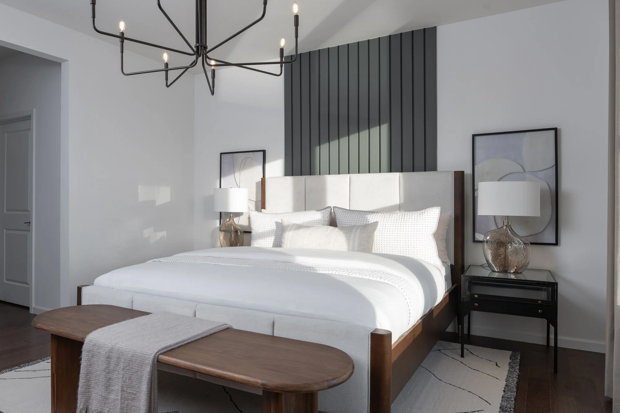 Modern bedroom with white bed, decorative pillows, black nightstand with lamp, framed artwork, dark wood accents, and a black geometric chandelier.