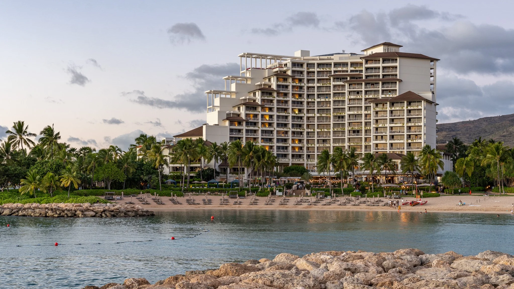 Four Seasons hotel with multiple floors, surrounded by palm trees, near the water with lounge chairs on the sandy beach and a rocky shoreline in the foreground.