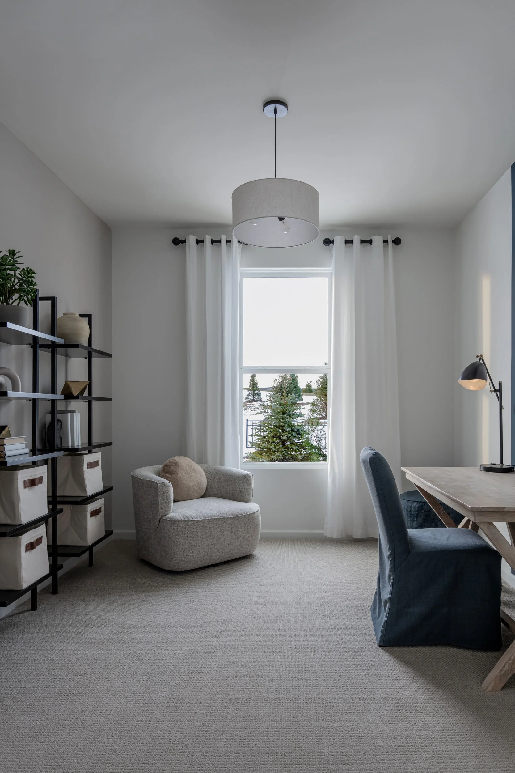 A bright room with a window showing snow-covered trees outdoors, white curtains, a beige armchair with a round cushion, a black bookshelf with decorative items, a wooden desk with a black desk lamp, and a gray upholstered chair.