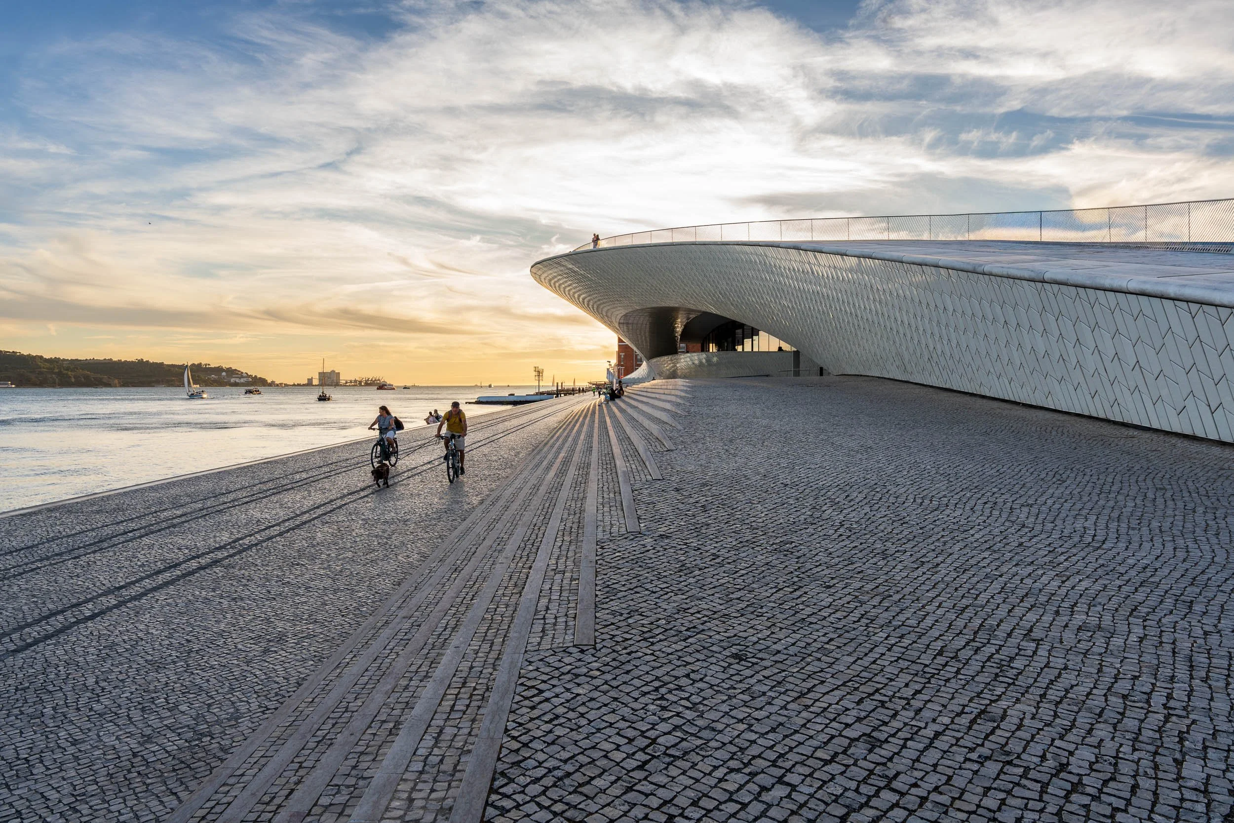 People riding bikes along waterfront promenade at sunset, modern building with curved architecture on the right, sailboats on the water, cloudy sky overhead.
