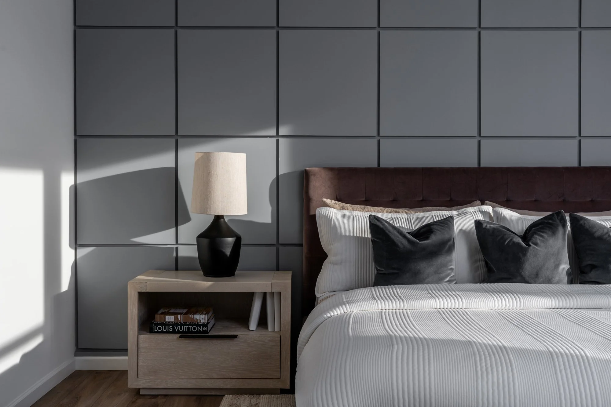 A modern bedroom with a beige nightstand, a black and beige table lamp, a bed with a brown upholstered headboard, white and gray pillows, and a gray padded wall behind the bed.