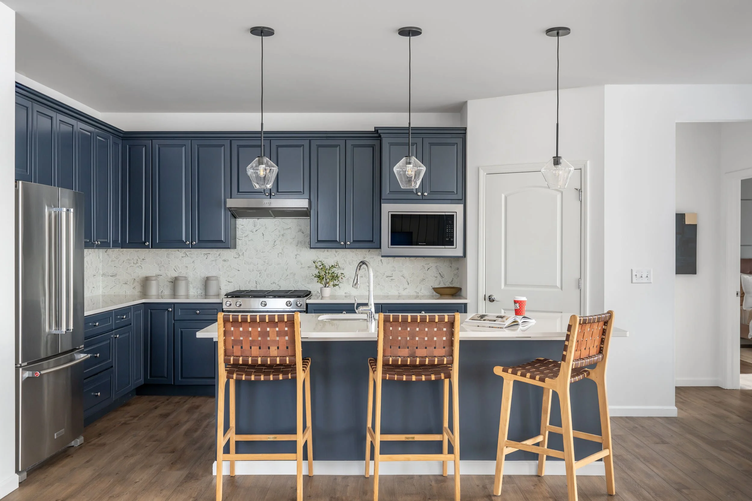 Modern kitchen with navy blue cabinets, a white island with a sink, three woven wood bar stools, and pendant lights, with a stainless steel refrigerator and a microwave.