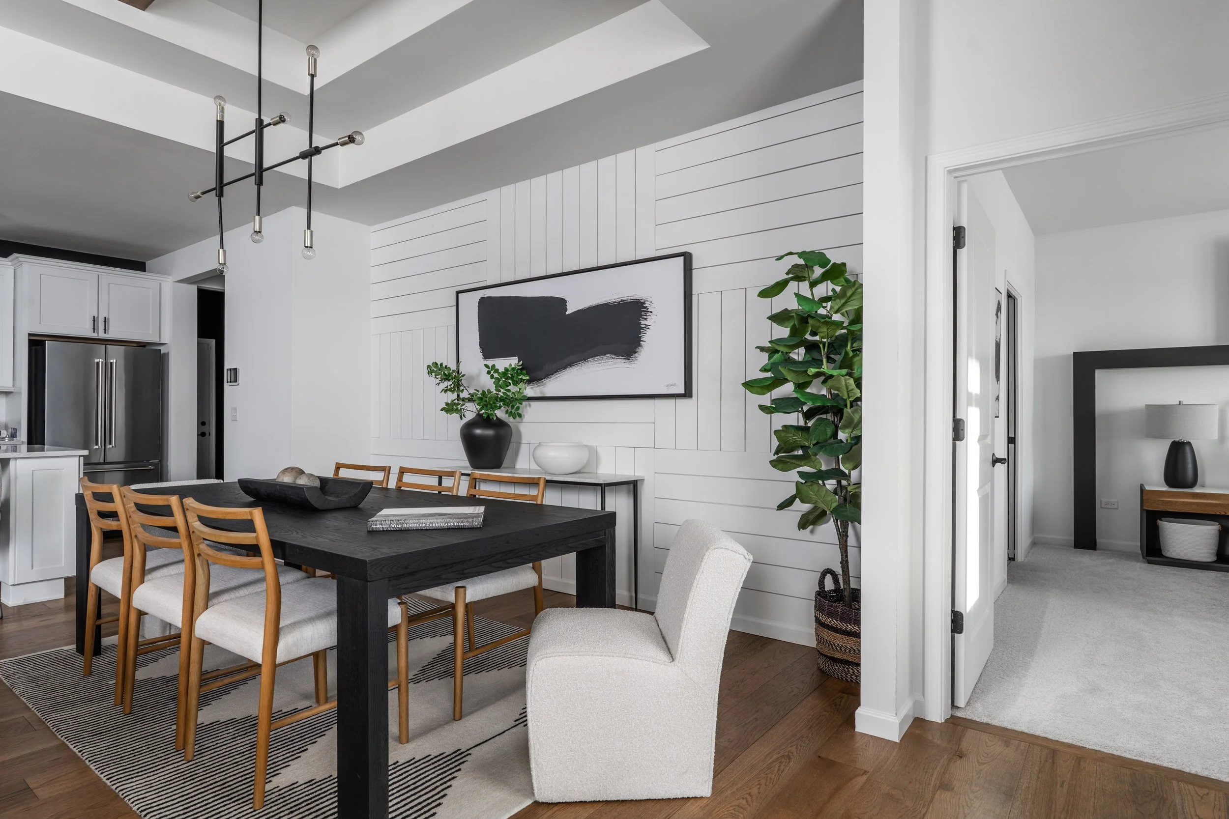 Modern dining room with a black wood table, six wooden chairs with white cushions, and one white armchair. White paneled wall with black and white abstract painting, large green potted plant, black vase, and white bowl surface. Adjacent kitchen with white cabinets and stainless steel refrigerator. Part of a cozy sitting room with a side table and lamp.