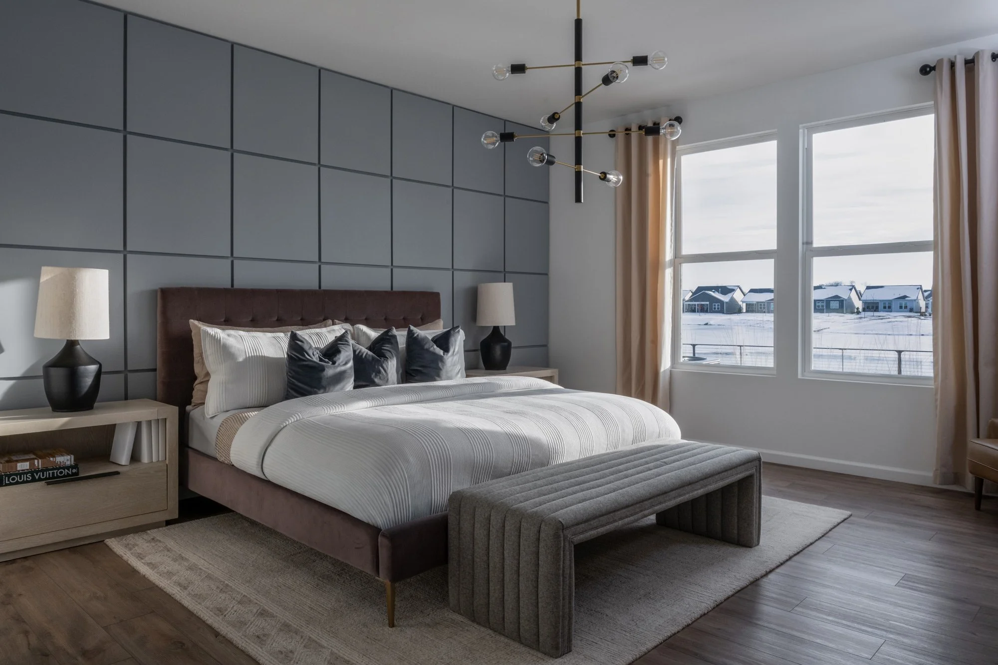 Modern bedroom with large windows showing a snowy landscape and houses outside. The room features a bed with a tufted headboard, gray pillows, and a striped duvet. A bench sits at the foot of the bed, and two nightstands with lamps are on each side. 