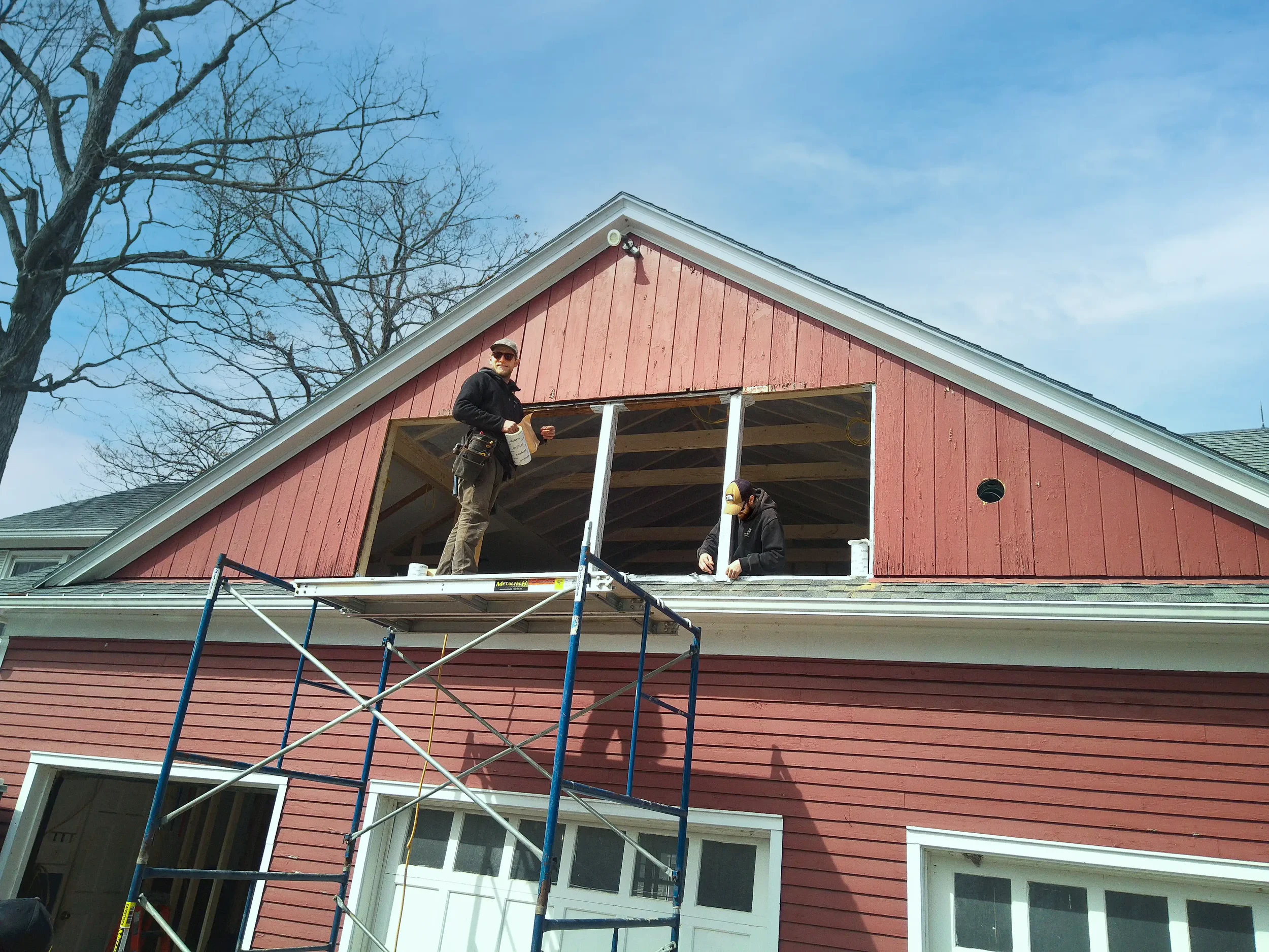 Two carpenters on scaffolding installing a large window in the upper story of a house painted red with white trim.