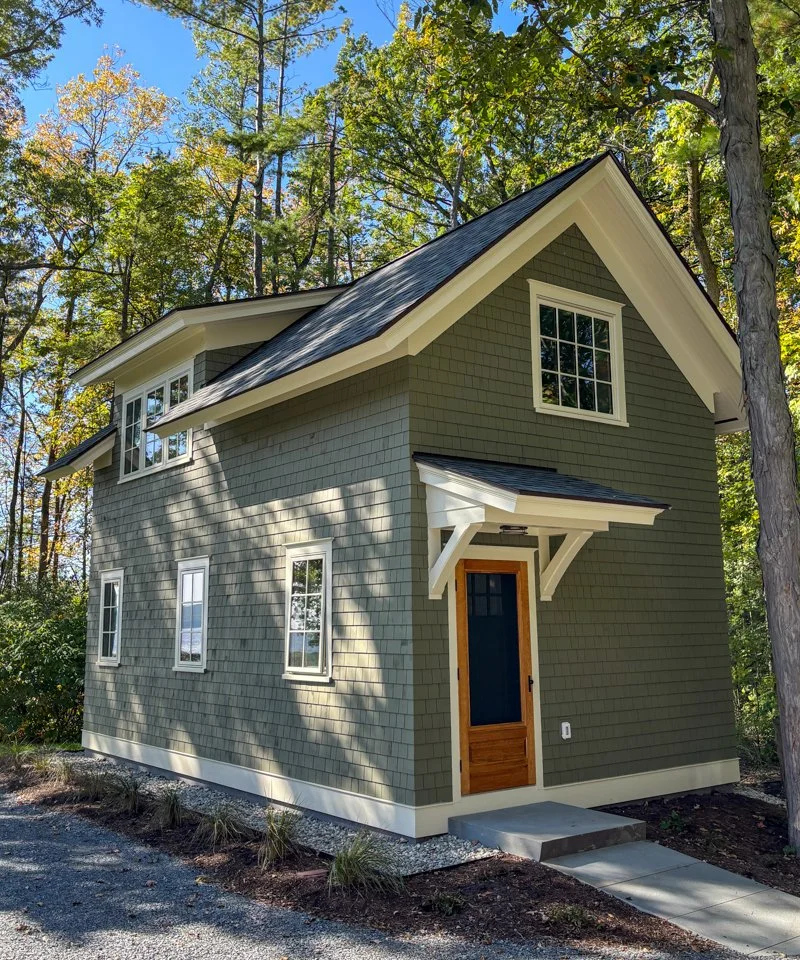 Small house with sage green shingled siding, cream trim, and a natural wood door. Behind the house are trees and blue sky.