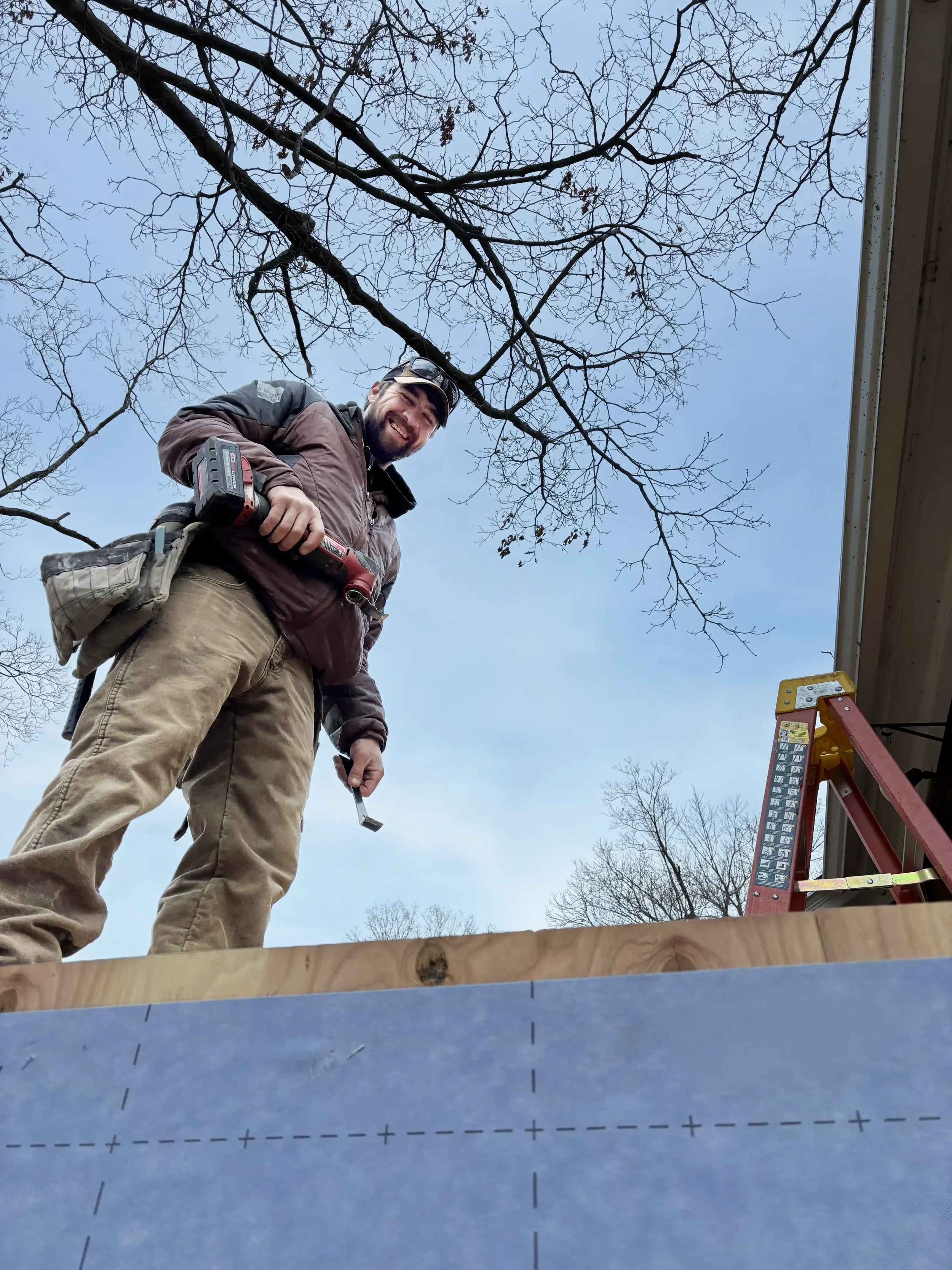 A carpenter standing outdoors on a construction site, smiling and holding a power drill.