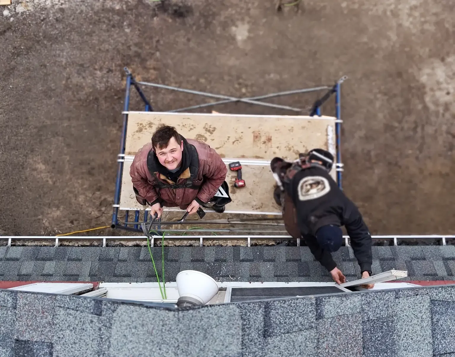 Two carpenters on a roof. One is smiling and looking up at the camera, wearing a brown jacket and black gloves. The other worker, in black safety gear, is reaching toward the edge of the roof.