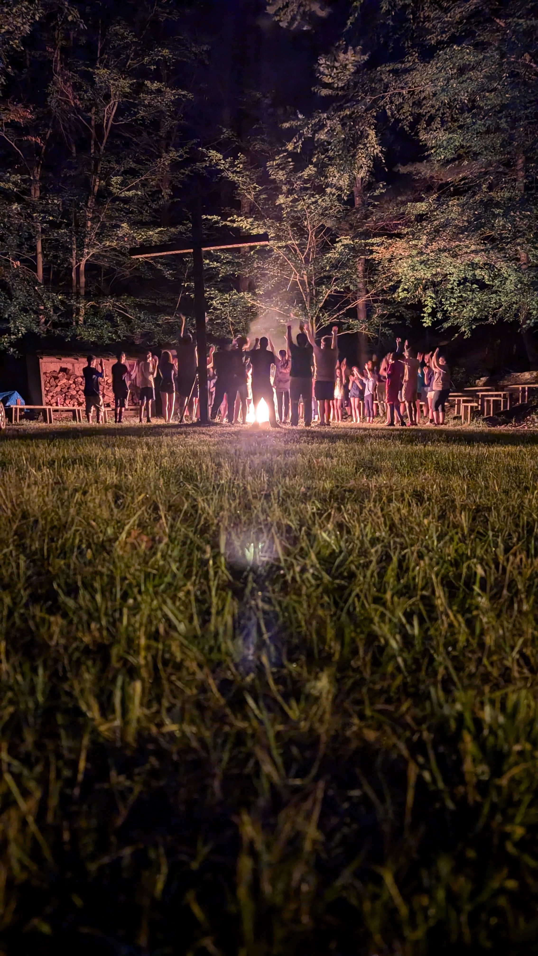 a view of grass leading to students surrounded by a campfire singing and dancing