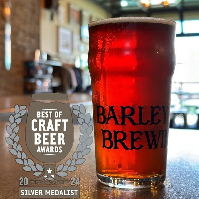 A glass of amber-colored beer with a foamy head on top, with a 'Barley Brew' label, placed inside a brewery or pub setting with blurred background. An award medal for 'Best of Craft Beer Awards' from 2024, Silver Medalist, is next to the glass.