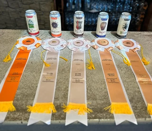 Five canned beverages placed behind five award ribbons on a gray surface. The ribbons are orange, white, white with silver, orange with bronze, and white with gold, each indicating different awards at an event in 2024.