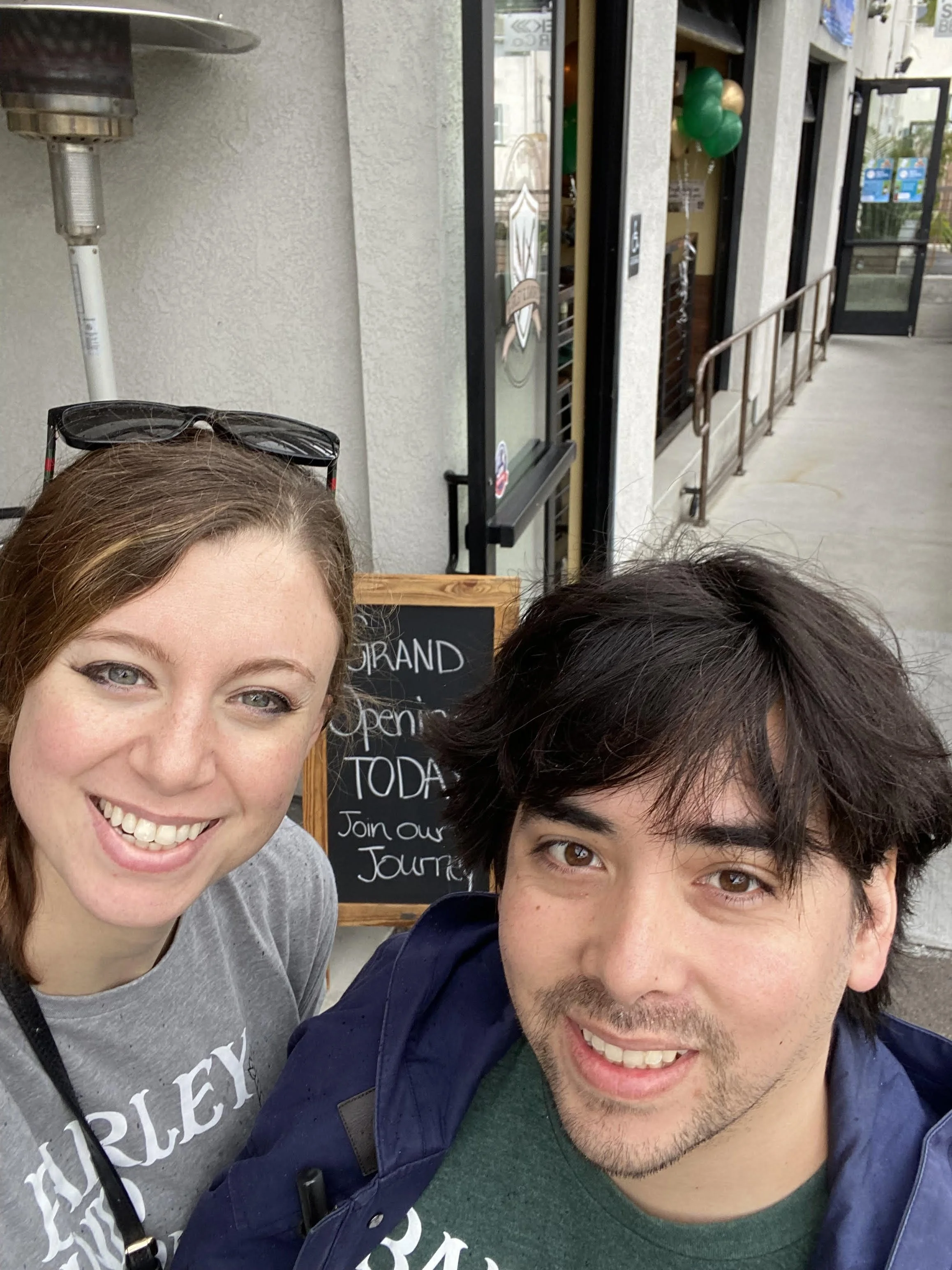 A smiling woman with sunglasses on her head and a man with dark hair and facial hair taking a selfie outside a storefront with a chalkboard sign.