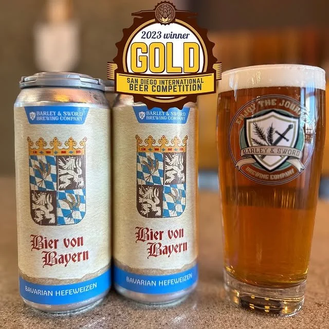 Two cans of Bavaria Hefeweizen beer and a pint glass filled with beer, displaying the Barley & Sword Brewing Company logo, with a gold award badge at the top indicating 2023 Gold winner at the San Diego International Beer Competition.