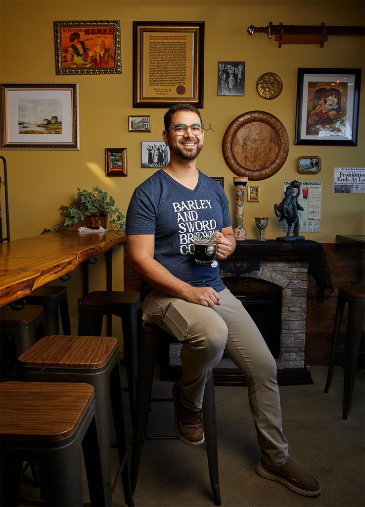 Man smiling and sitting on a barstool in a decorated room, holding a beer mug. The room has framed pictures and art on the wall, a fireplace with decorations, and a wooden bar counter.