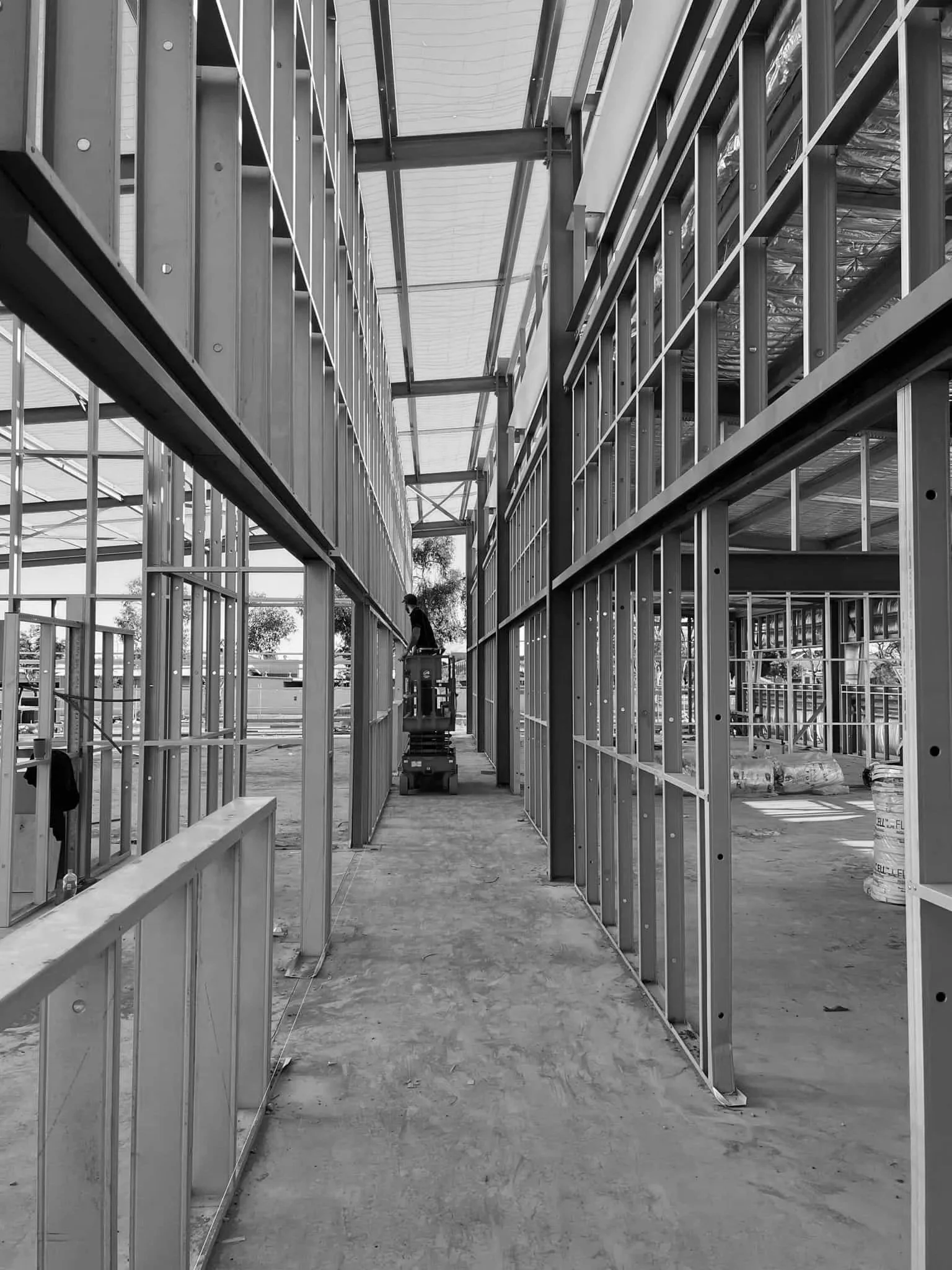 Construction site with metal framing for walls inside a building, with a worker on a lift working on the upper sections of the structure.