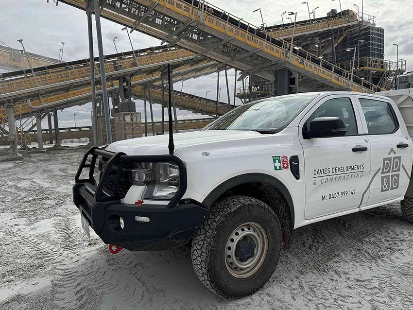 A white pickup truck with a black front bumper and grille, parked on a gravel surface, with an industrial conveyor system and a large building under construction in the background.