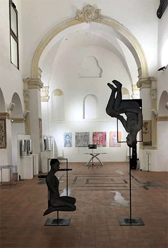 An art installation of two life-sized sculptures of people performing acrobatics, one sitting and the other upside down on stilts, inside a spacious white gallery with arches and abstract artwork on the walls.