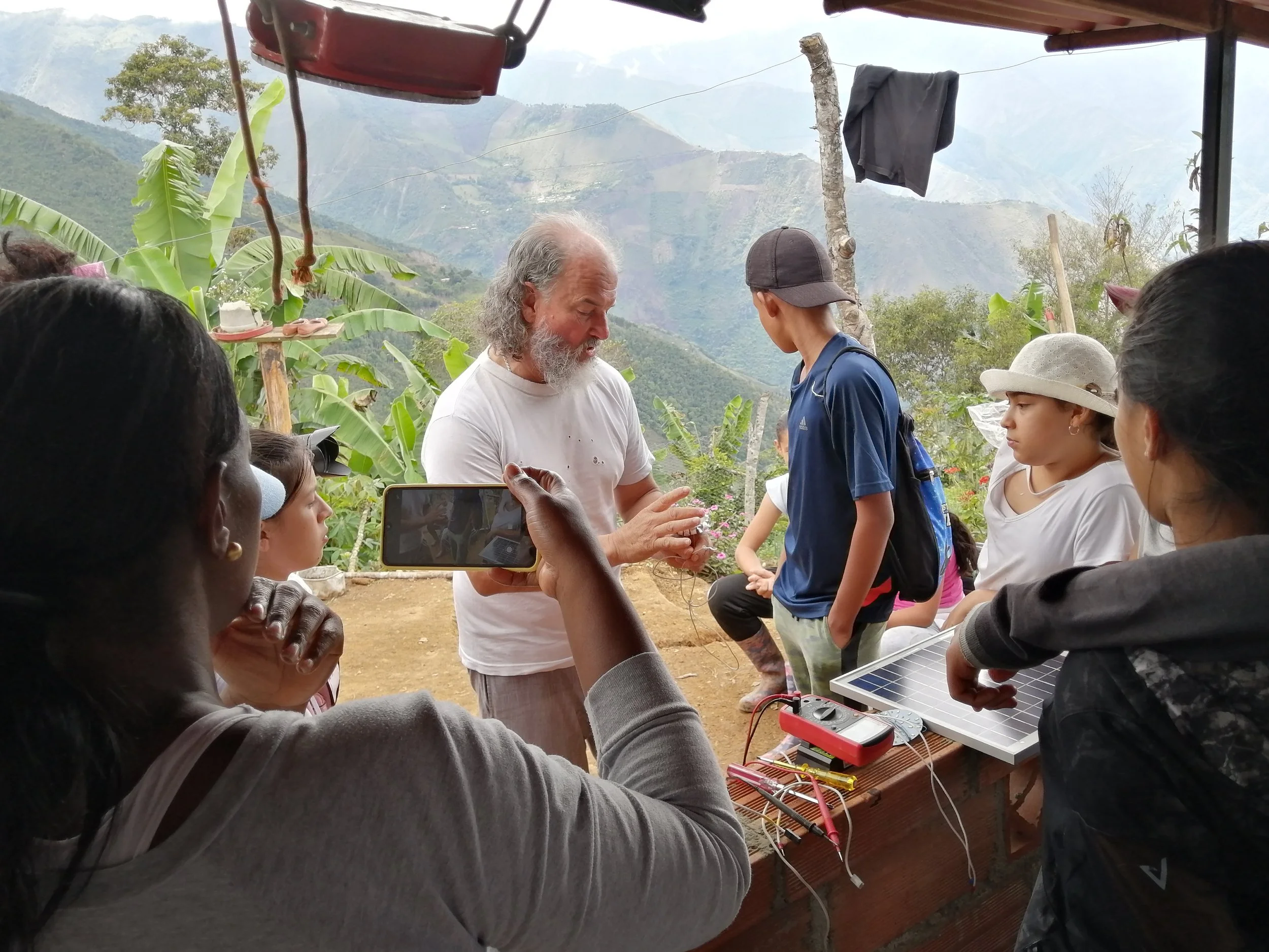 A group of children and adults gathered outdoors on a hillside, listening to an older man with white hair and a beard who is demonstrating something with his hands. Some people are taking pictures, and there is a small solar panel and electronic devices on a wooden table in the foreground. The background features lush green mountains and banana plants.