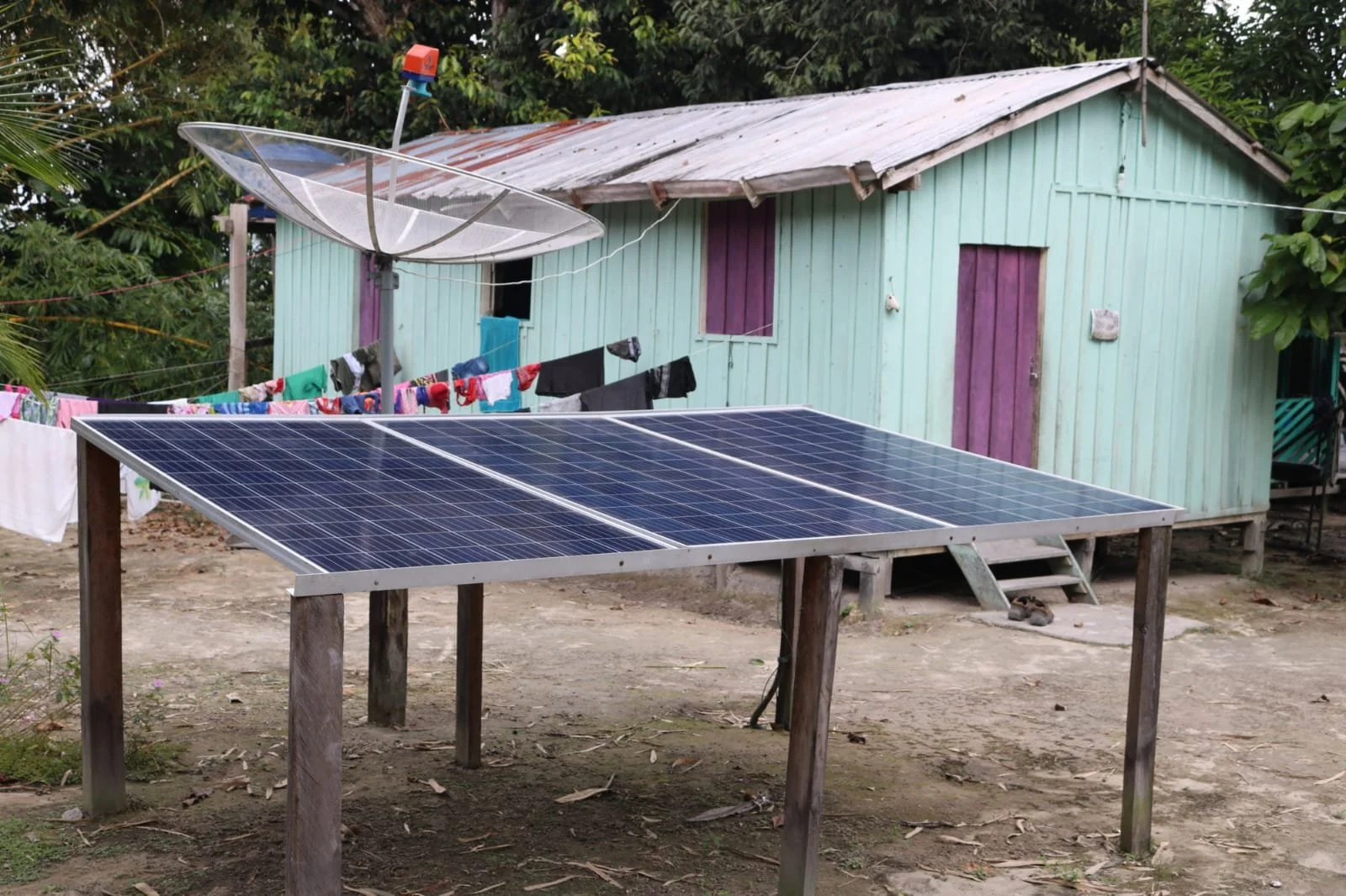 A small wooden house with light blue walls and purple shutters, with solar panels set up on raised wooden platforms in the foreground. Clothes are hanging on a clothesline near the house, and a satellite dish and solar-powered light are visible.