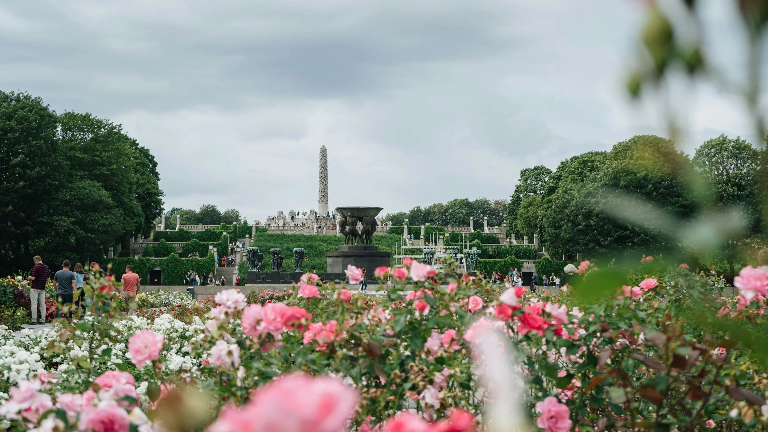 A lush park with pink and white flowers, featuring a stone monument surrounded by greenery, trees, and people walking.