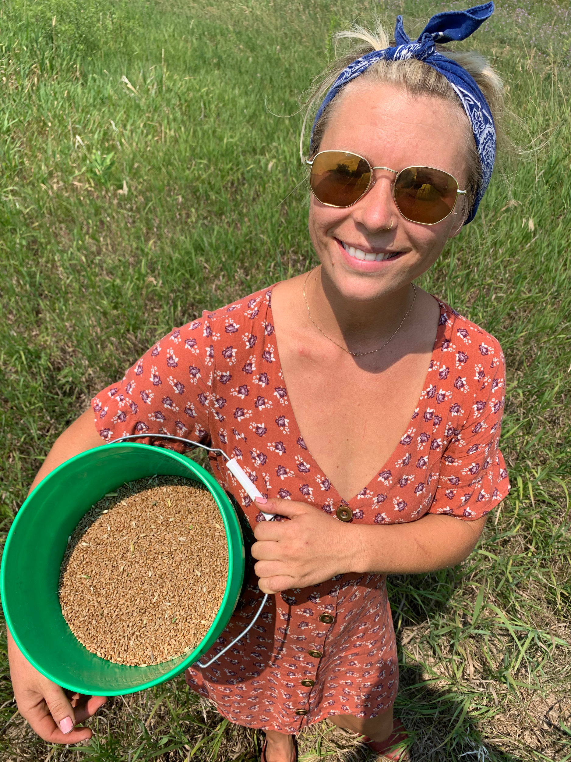 Person smiling outdoors, wearing sunglasses and a patterned dress, holding a green bucket filled with grains.