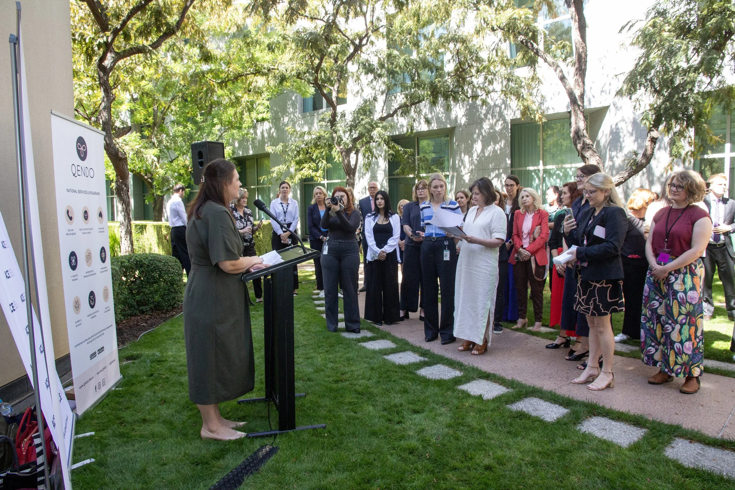 A woman in a green dress is speaking at a podium while an audience of diverse professionals listens outdoors in a green courtyard with trees and buildings in the background.