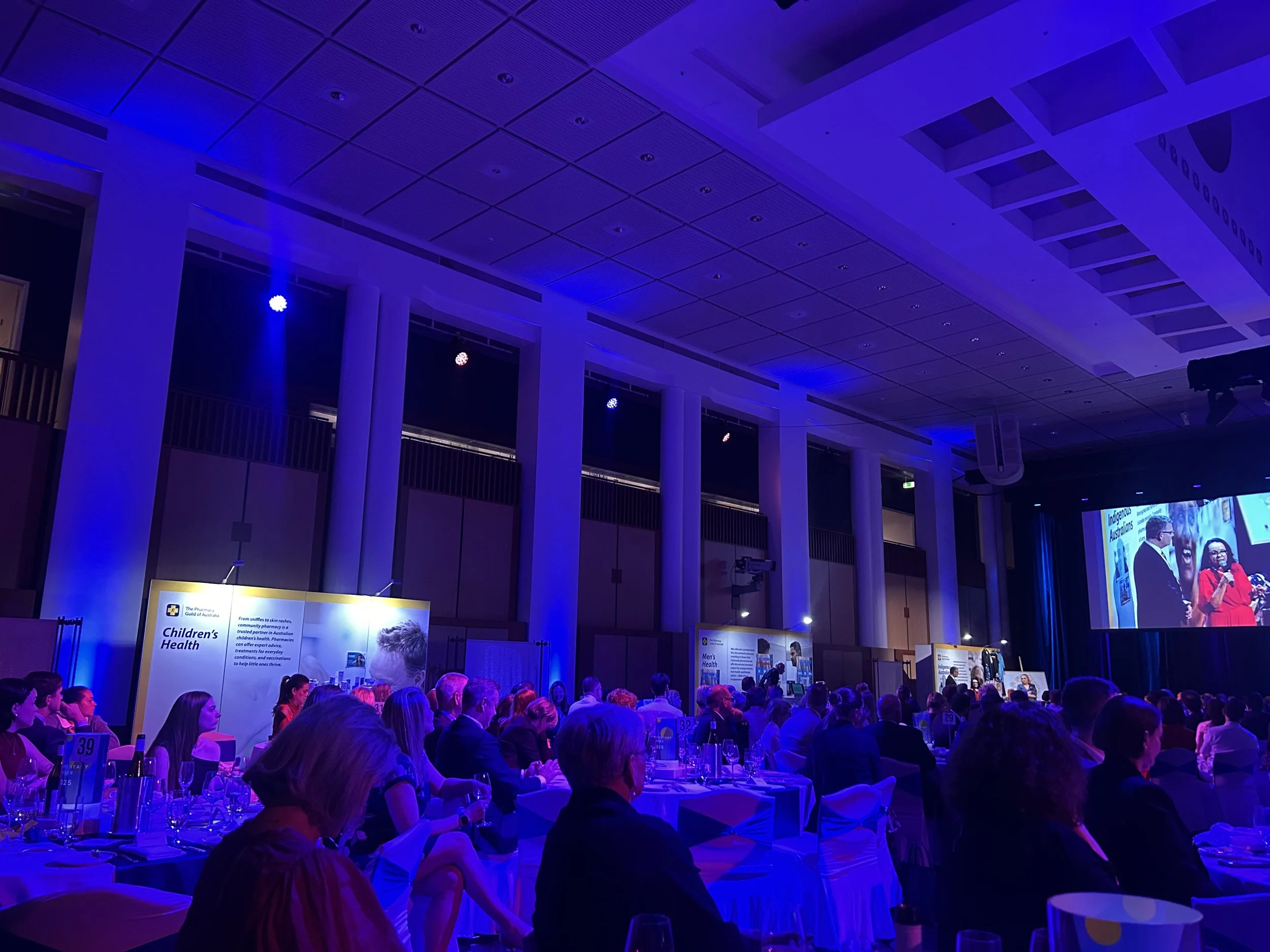 People attending a formal event or conference in a large, illuminated indoor hall with blue lighting, average-sized round tables with white tablecloths, and a stage with a large screen displaying a presentation or speech.