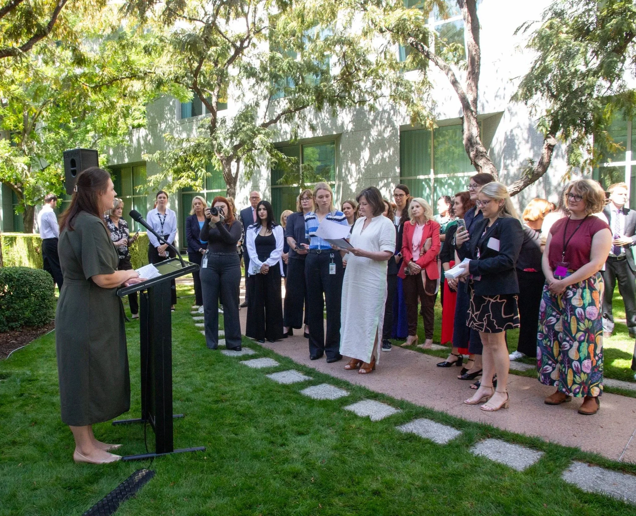 A woman in a green dress speaking at a podium to a group of people outdoors, gathered on a grassy area with trees and a building in the background.