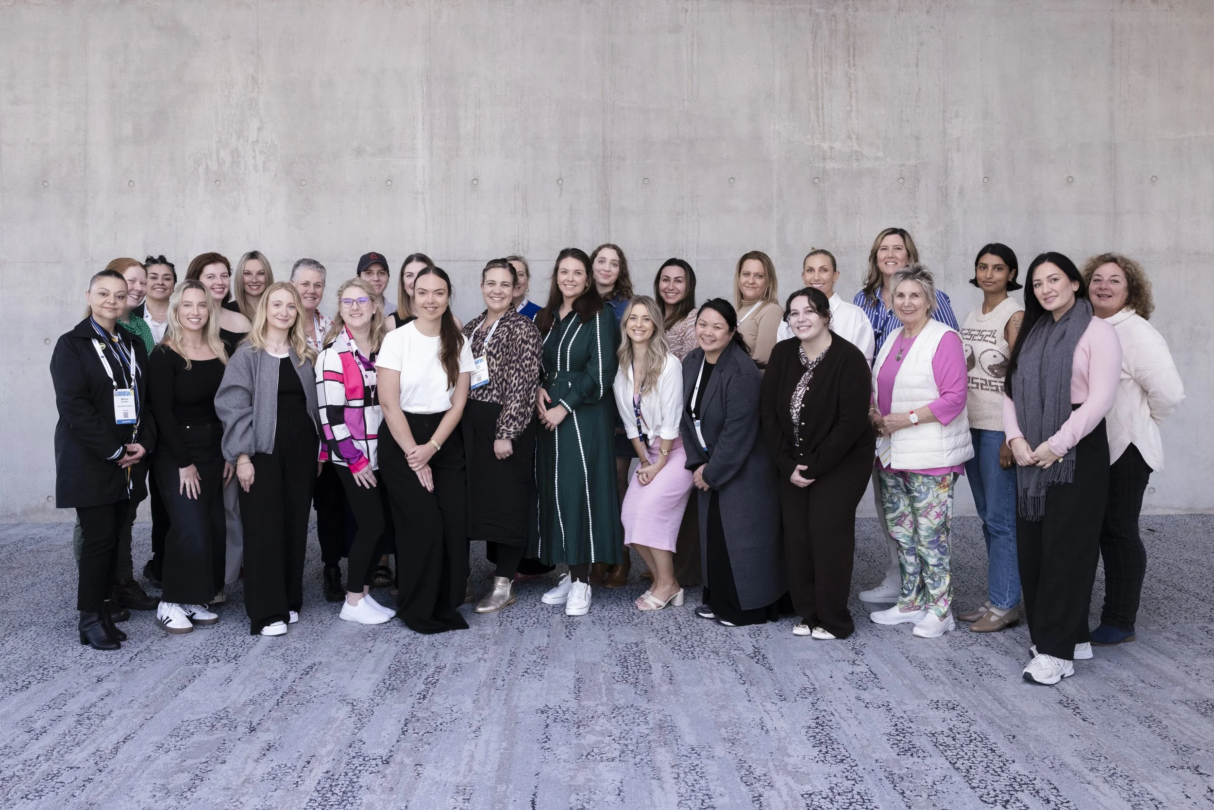 Group of women and men standing together, posing for a photo indoors against a plain wall.