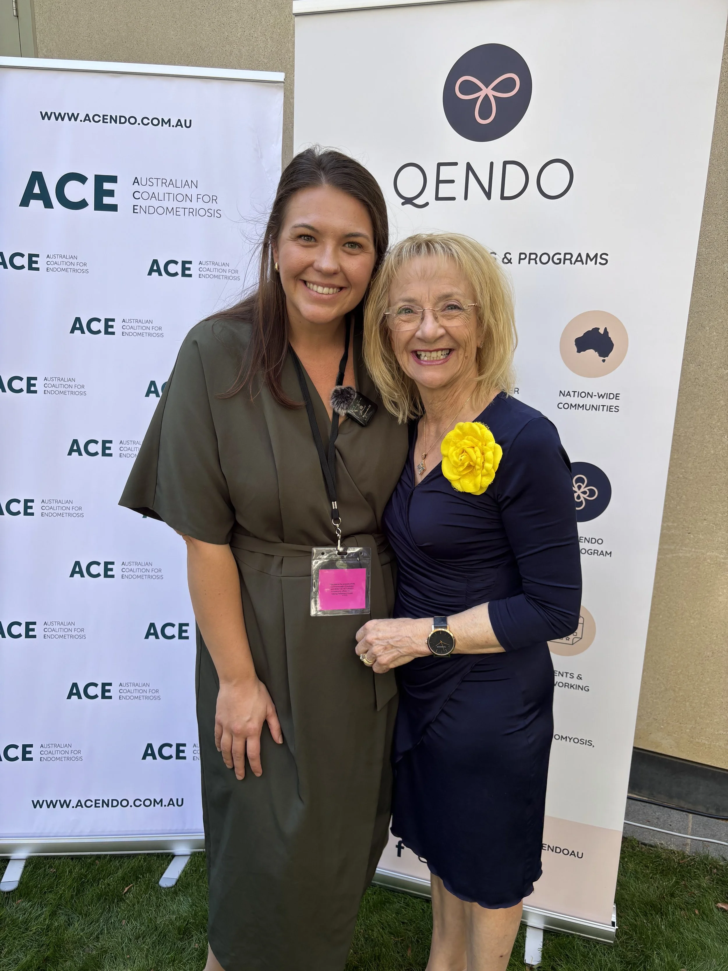 Two women smiling and standing close together outdoors in front of banners for ACE and QENDO, with one woman wearing a yellow flower pin and the other a badge.