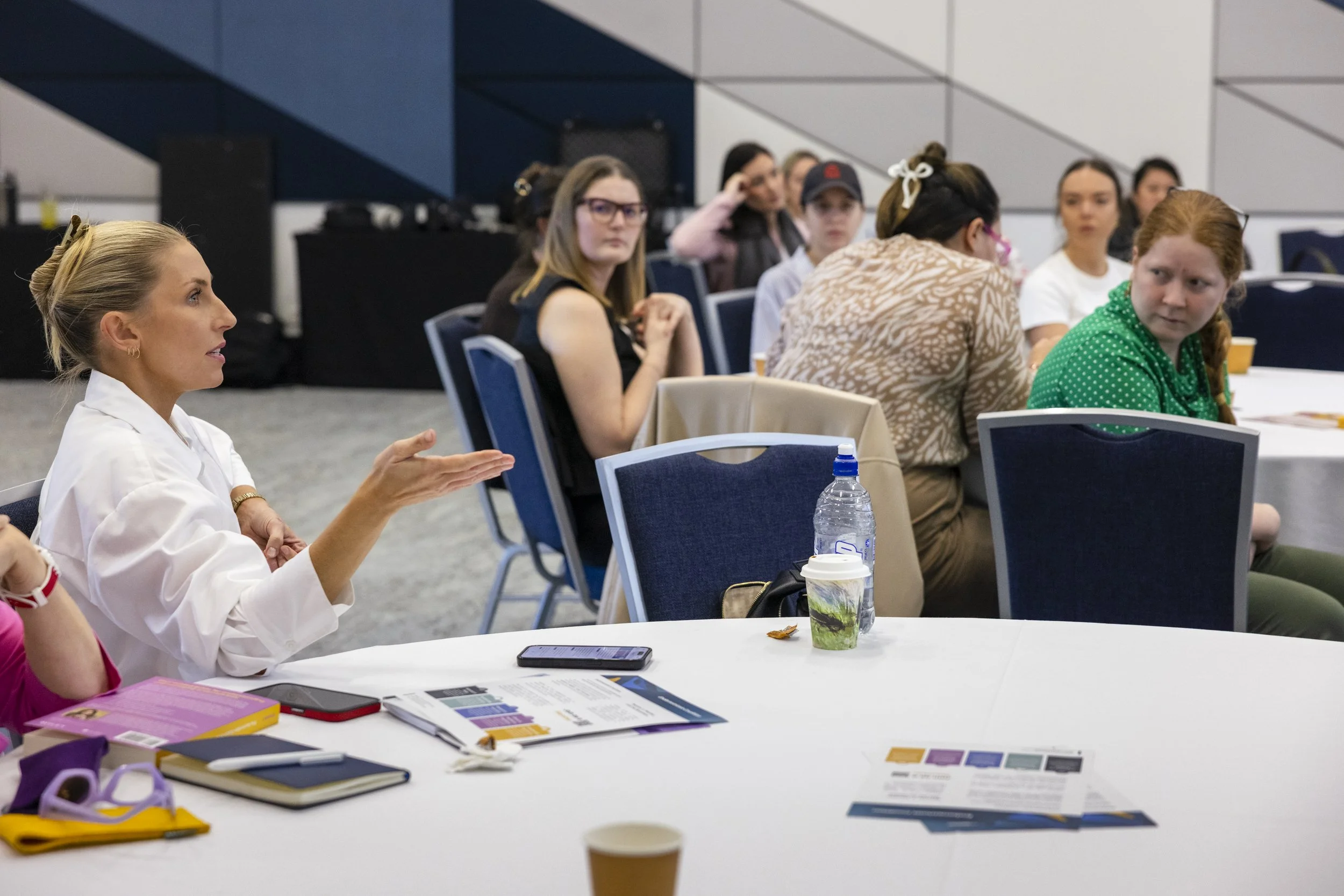 A woman in a white shirt is speaking and gesturing with her hand at a round table with multiple women attending a meeting or conference in a room with modern decor.