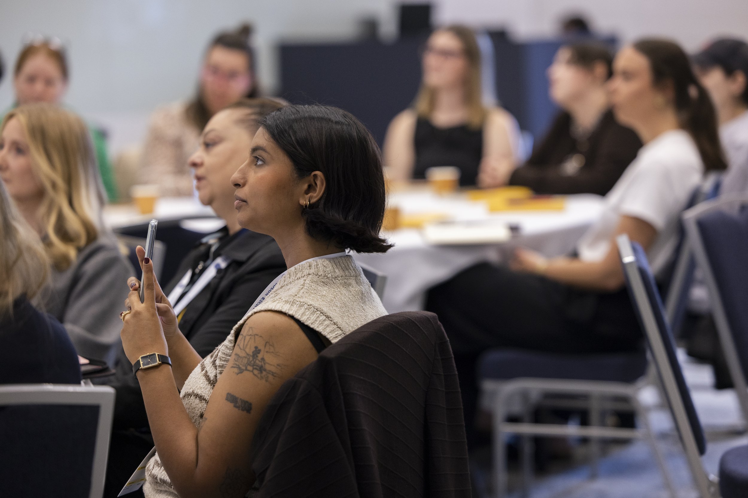 Women attending a conference or seminar, sitting at tables, with some taking notes and listening attentively.