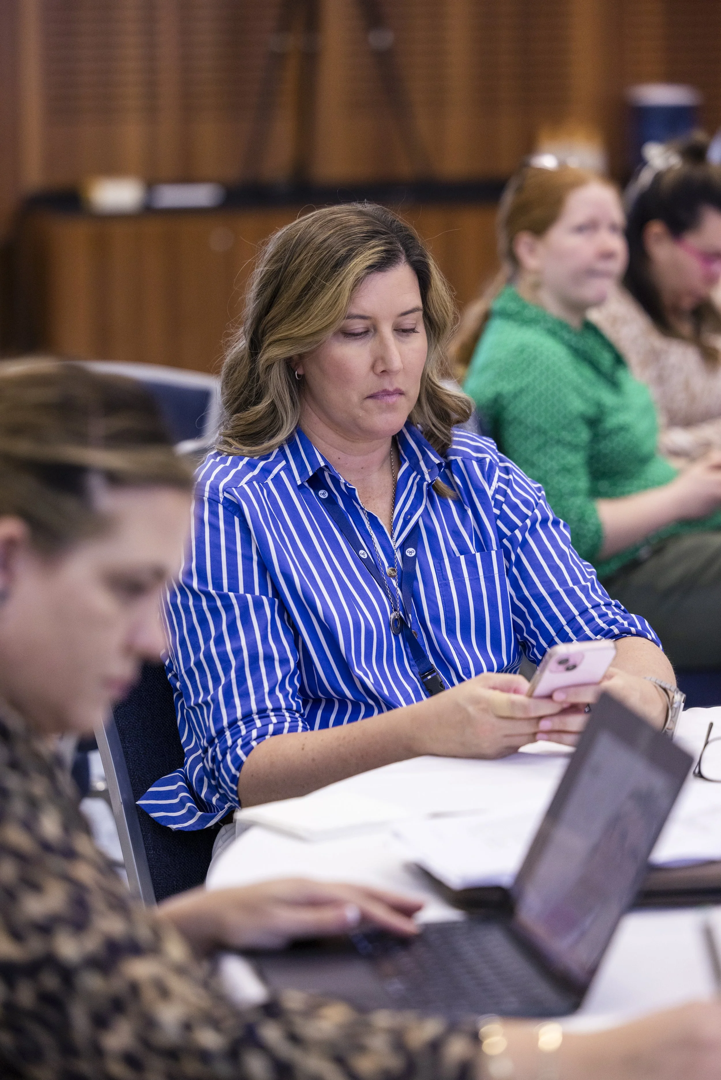 A woman in a blue and white striped shirt sitting at a table with a laptop and a smartphone, focusing on her phone, in a meeting or conference setting.