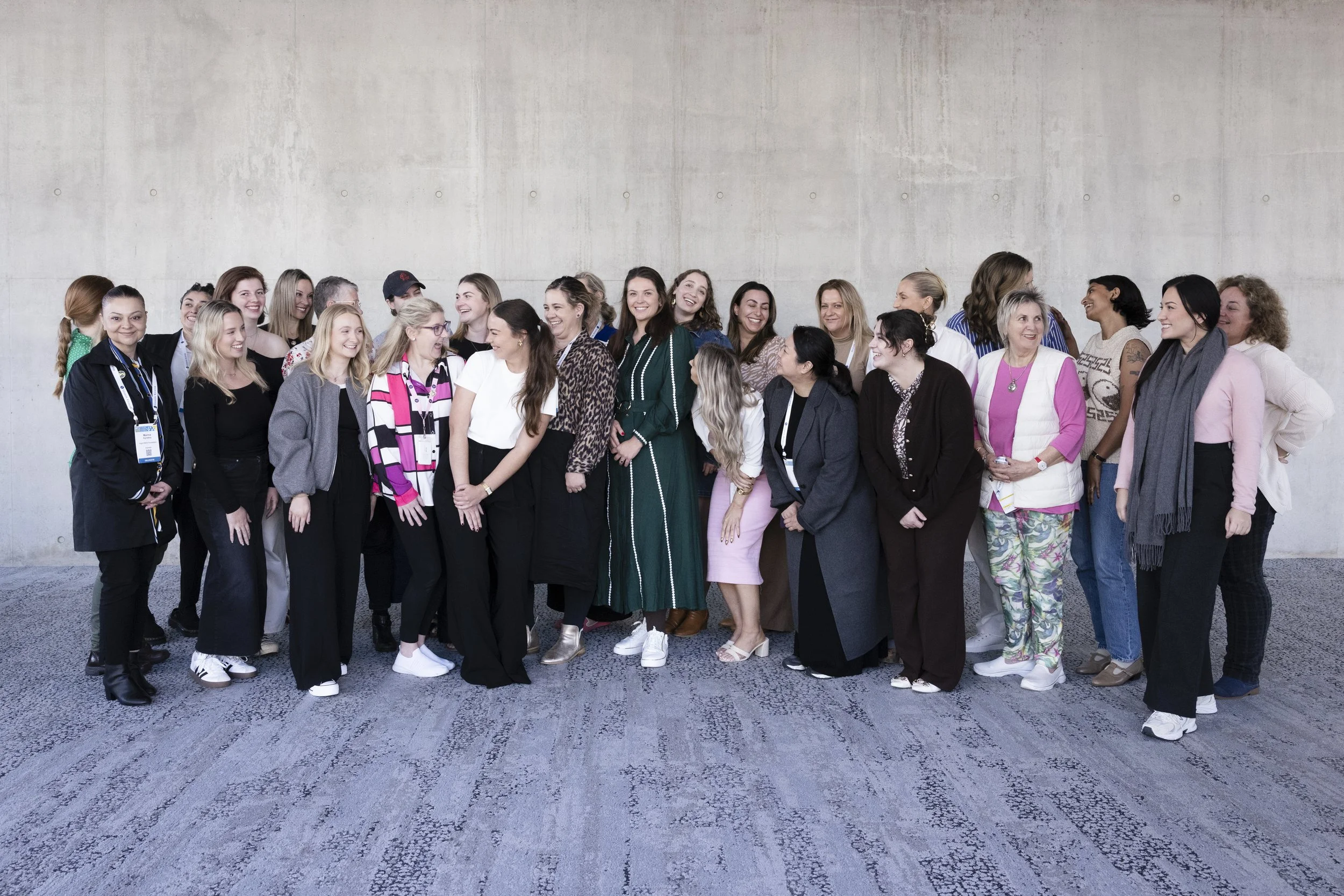 Group of diverse women standing together indoors with a plain concrete wall background, smiling and talking.