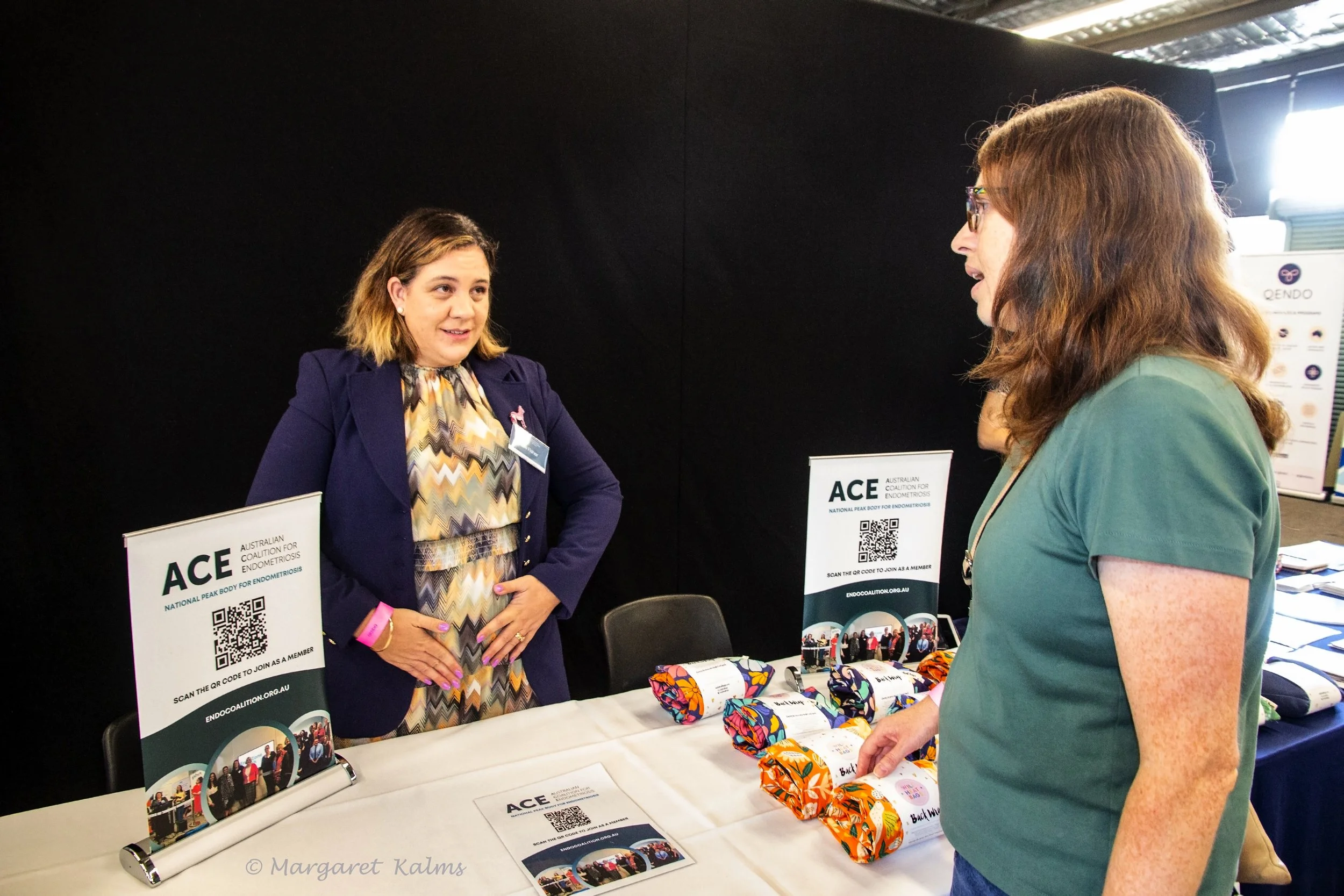 Two women conversing at a booth promoting Endometriosis awareness, with displayed informational signs, brochures, and colorful fabric items on the table.