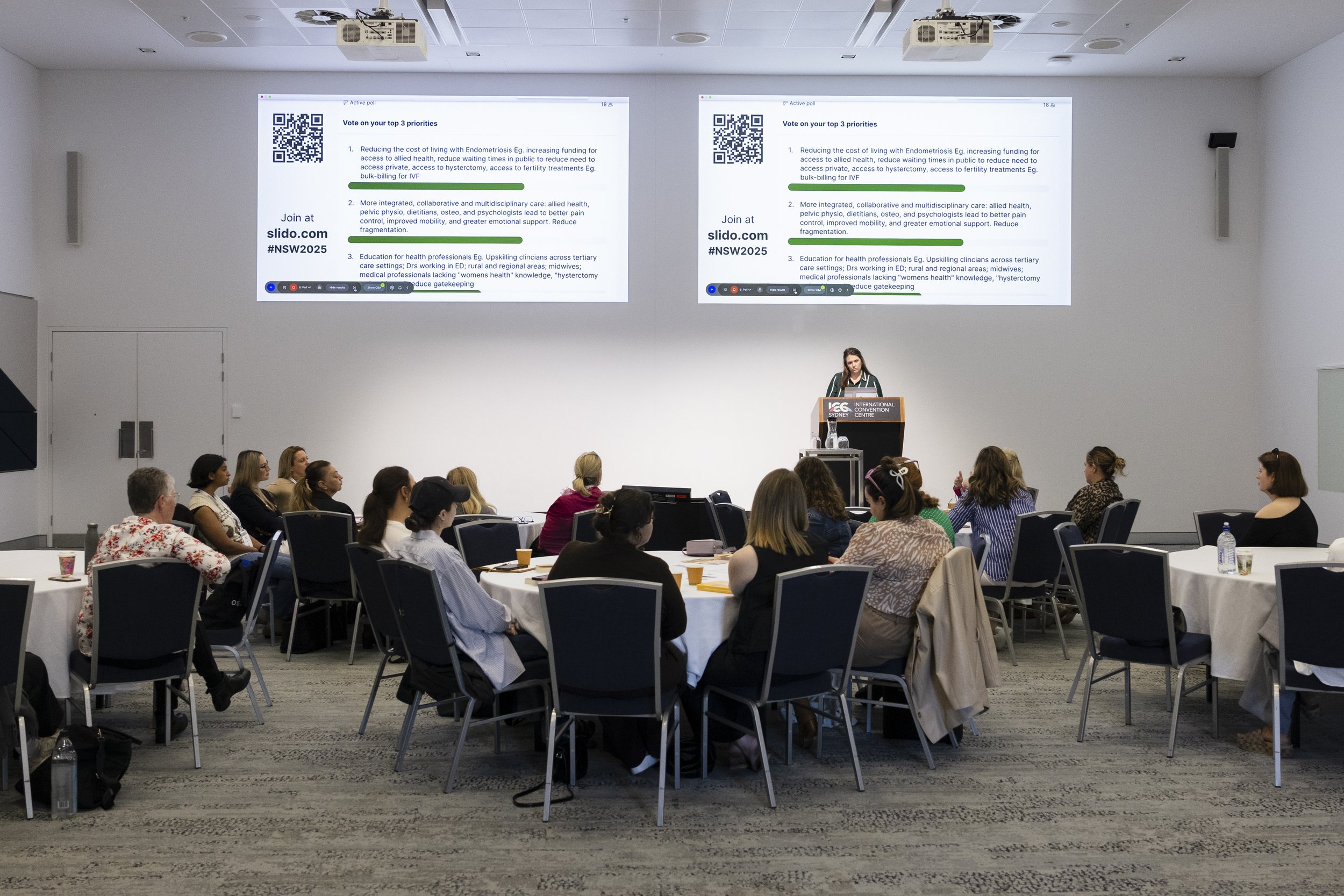 A woman presenting at a conference with two large screens displaying a slide behind her. The slide has a QR code and text about voting on top priorities. Audience members are seated at round tables, listening attentively in a bright, modern conference room.