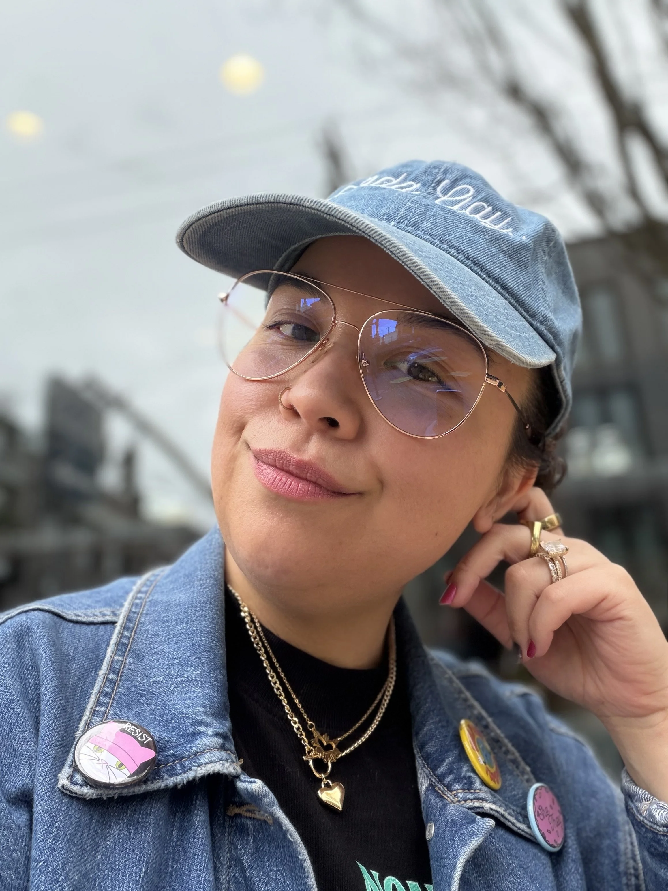 Angela is smiling, they are wearing large wire rimmed glasses, a denim hat that says "kinda gay", several gold chains, and a denim jacket with buttons.