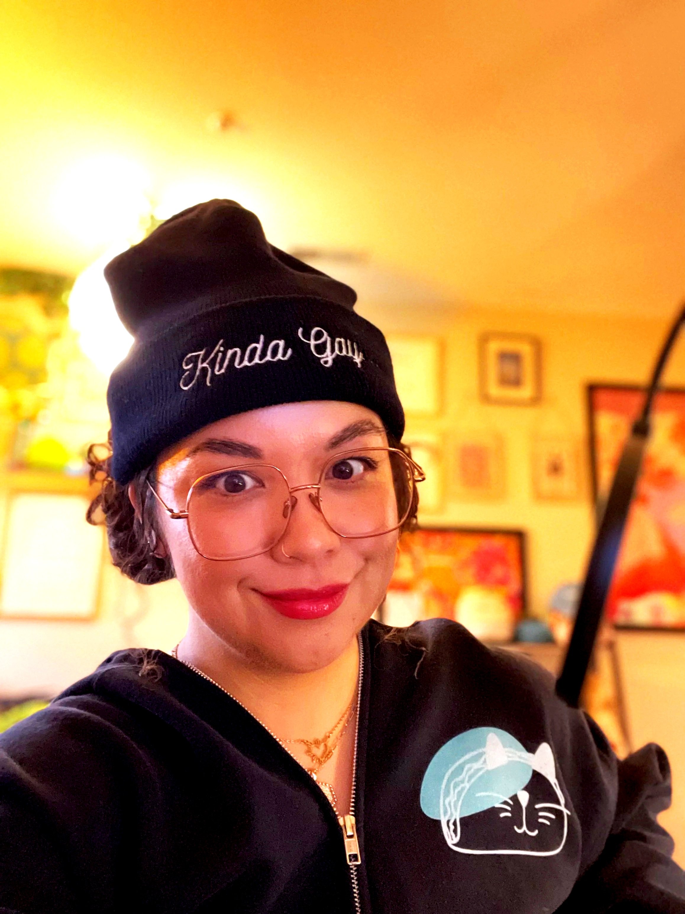 Angela is smiling. They're wearing a black beanie with "kinda gay" stitched on the front and a black hoodie with a teal Tacocat logo.