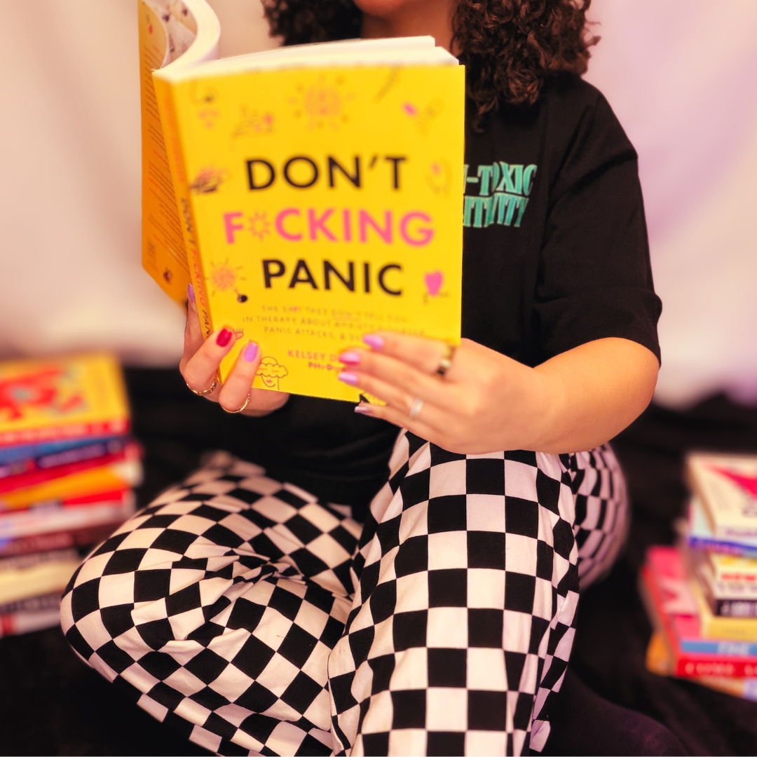 Angela is sitting and wearing black and white checkerboard pants. They're holding a bright yellow book that reads "Don't fucking panic"